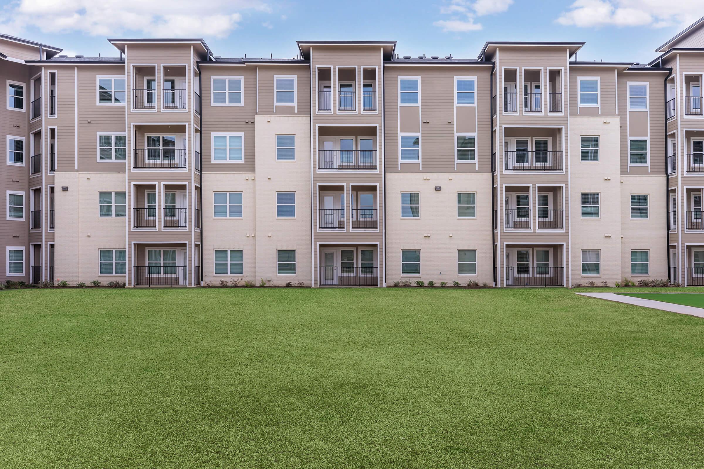 Modern apartment building with multiple floors featuring large windows and balconies. The structure is light-colored with a green lawn in the foreground, providing a spacious outdoor area. Clear blue sky above complements the contemporary architecture.