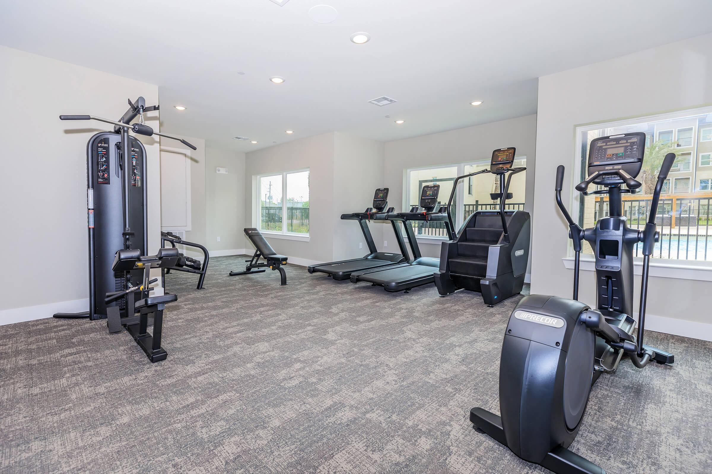 A modern fitness room featuring various exercise equipment, including a treadmill, elliptical machine, strength training machine, and a weight bench. Large windows allow natural light to brighten the space, and the flooring is carpeted for added comfort.