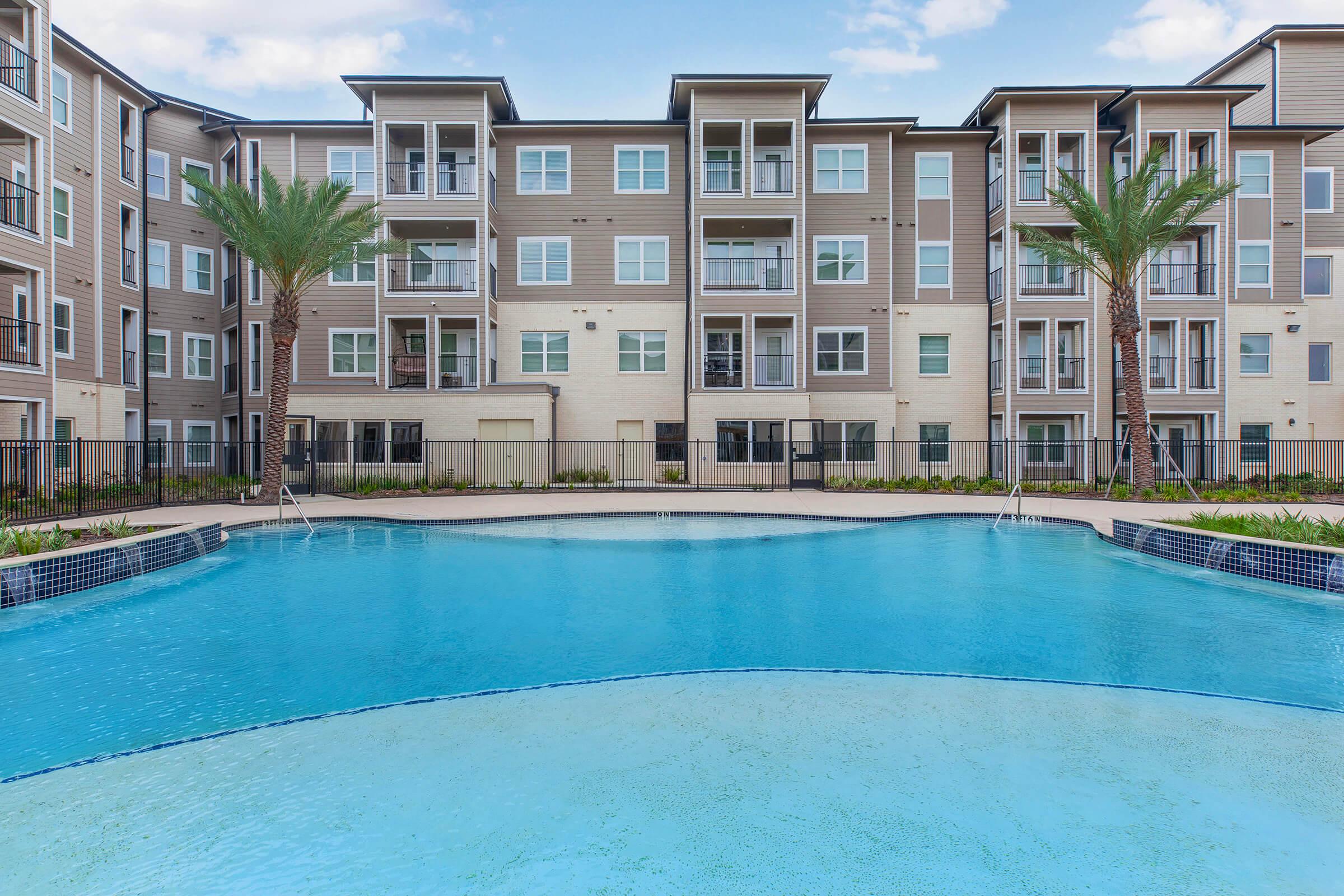 A large swimming pool with a curved edge in the foreground, surrounded by landscaped greenery. In the background, a modern multi-story apartment building with balconies and multiple large windows. Palm trees are integrated into the landscaping, creating a tropical feel. The sky is partly cloudy.