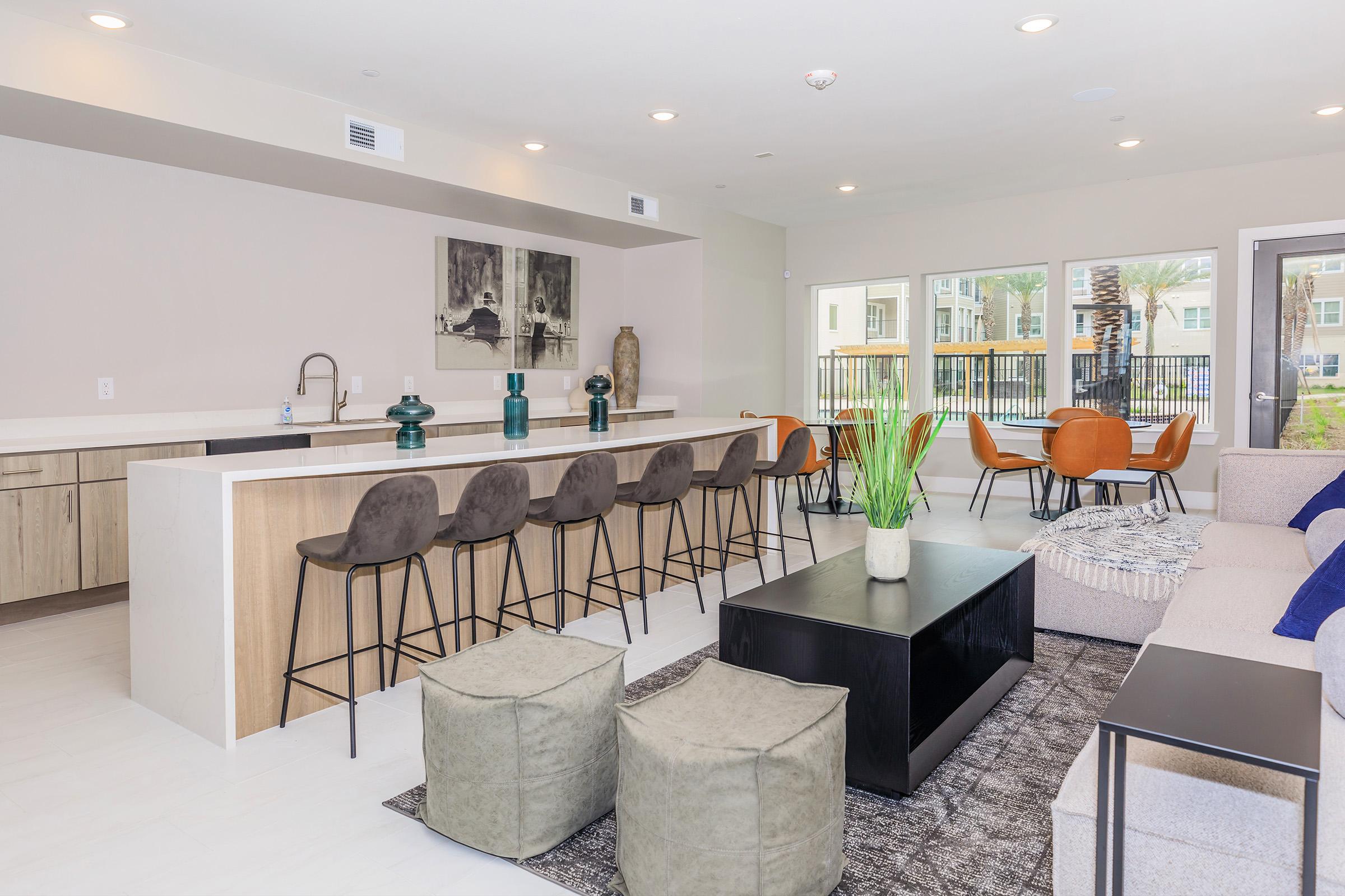 A modern living space featuring a stylish kitchen bar with high stools, a cozy seating area with a sofa and coffee table, and decorative plants. Large windows provide natural light, and there are chairs and artwork enhancing the ambiance, creating a welcoming atmosphere.
