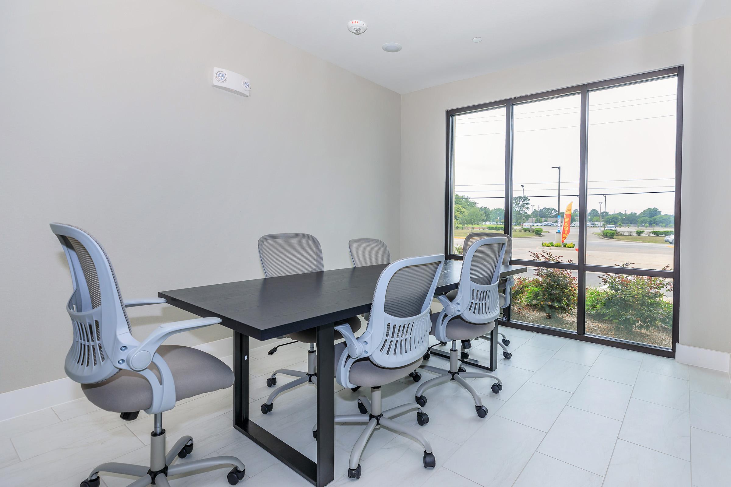 A modern conference room featuring a large black table surrounded by five ergonomic chairs. The room has large windows providing natural light and a view of an outdoor landscape with trees and parking area. The walls are painted in a light color, contributing to a spacious and inviting atmosphere.