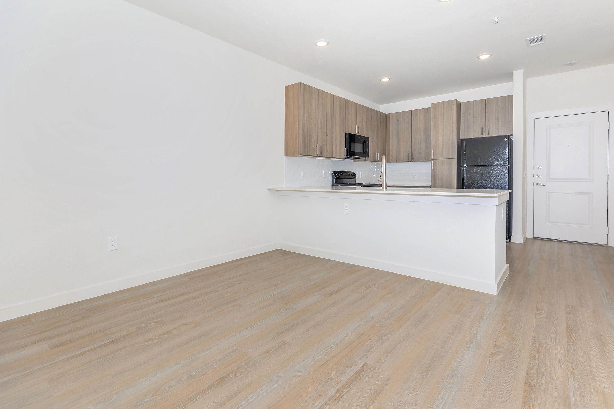 A modern, unfurnished kitchen and living area featuring light wood flooring, white walls, and a large island with a sink. The kitchen includes wooden cabinets, a microwave, and a black refrigerator. Natural light streams in, creating an airy atmosphere.