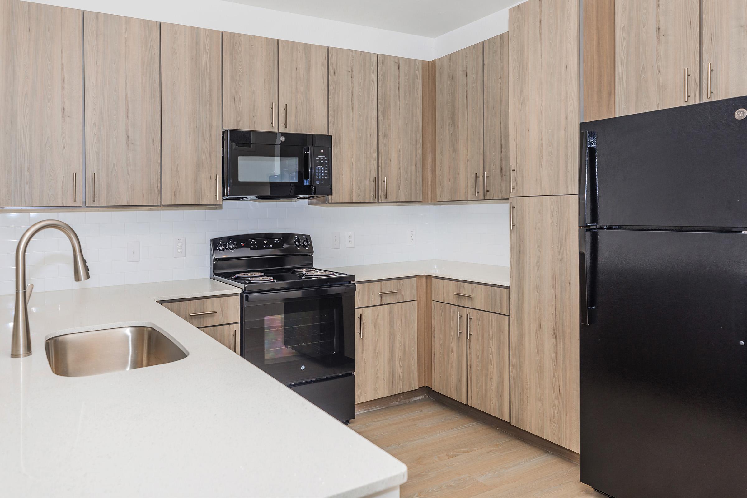 Modern kitchen featuring light wood cabinetry, stainless steel sink, black refrigerator, microwave, and oven. The countertops are white with a subtle texture, and the flooring is light wood. The layout is compact and functional, ideal for small spaces.