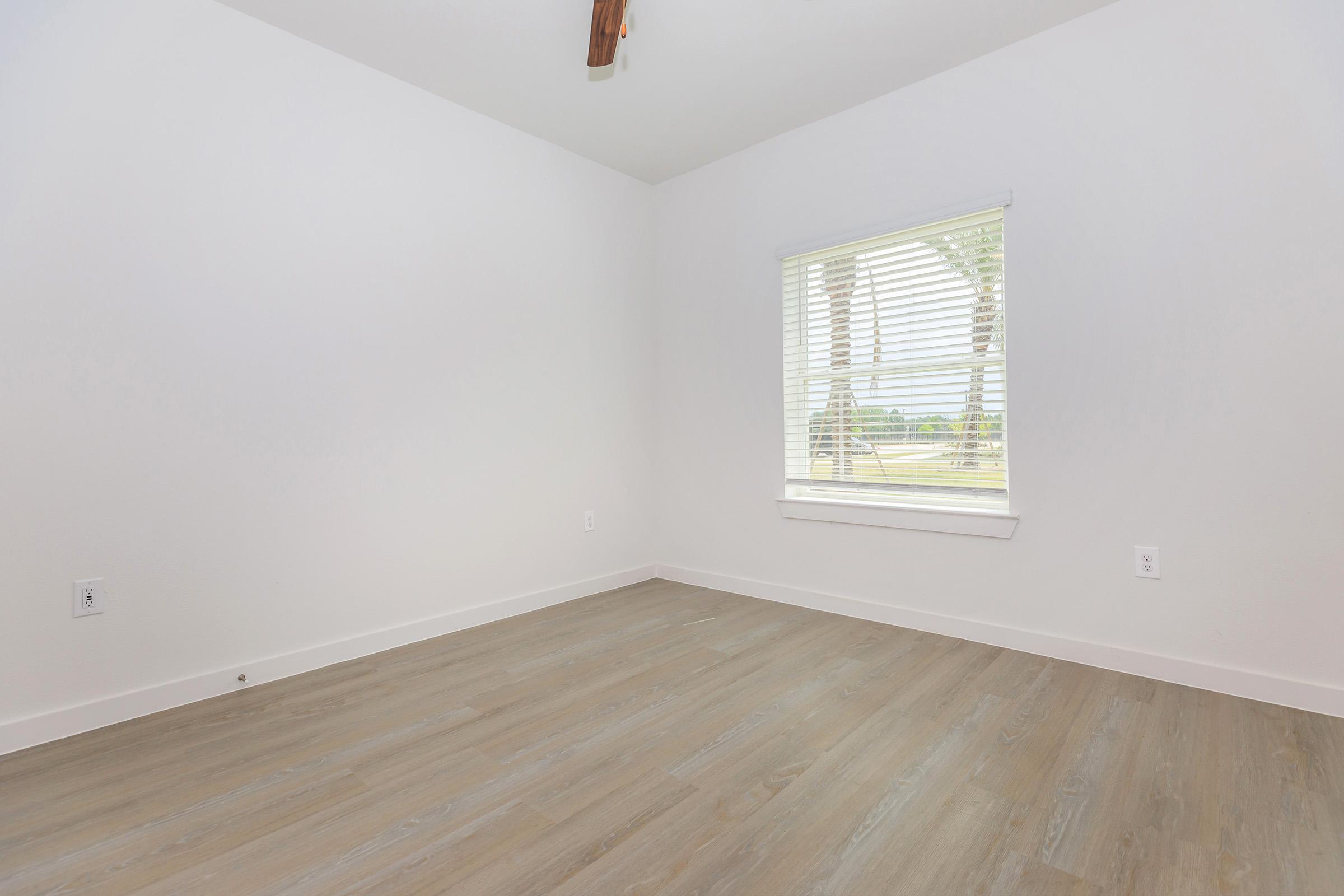 A bright, empty room with light-colored walls and a wooden floor. A window with white blinds allows natural light to enter, revealing a view of greenery outside. The ceiling features a ceiling fan, adding to the clean and simple design of the space.