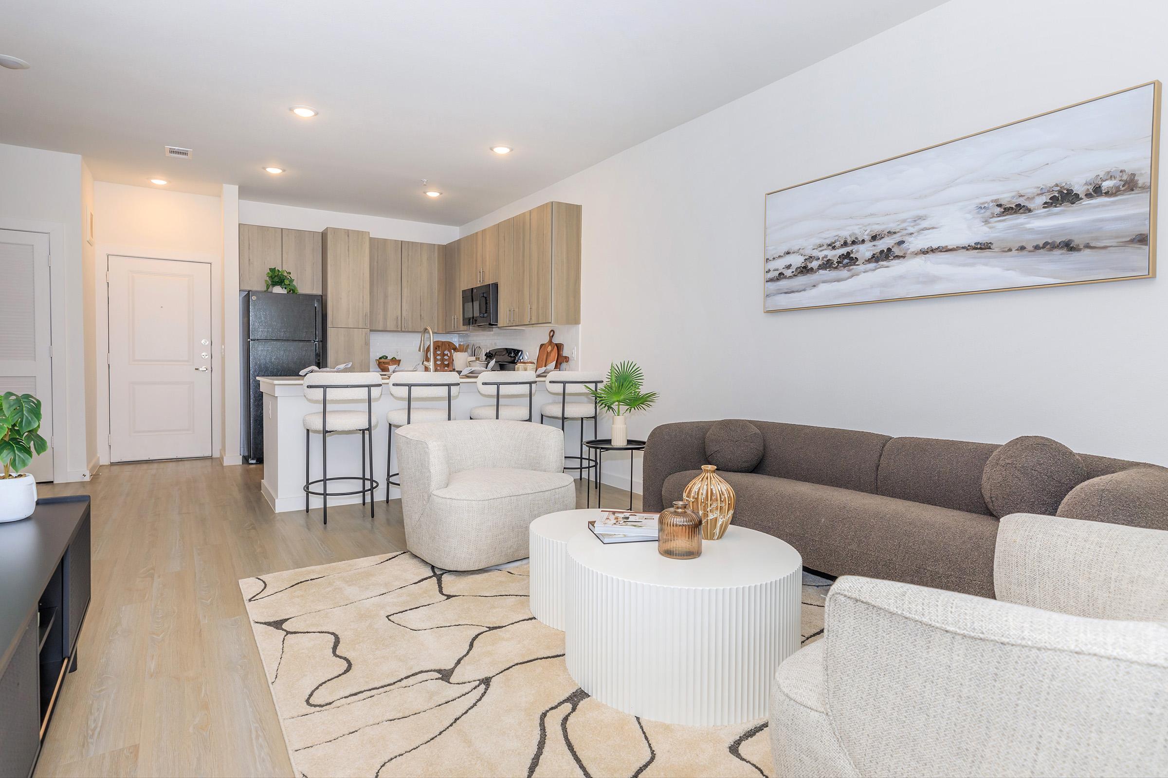 A modern living room featuring a cozy sofa and two armchairs surrounding a round coffee table. The space has light wood flooring and a neutral color palette, with a large painting on the wall. In the background, there's a kitchen area with bar stools and appliances. A small plant adds a touch of greenery.