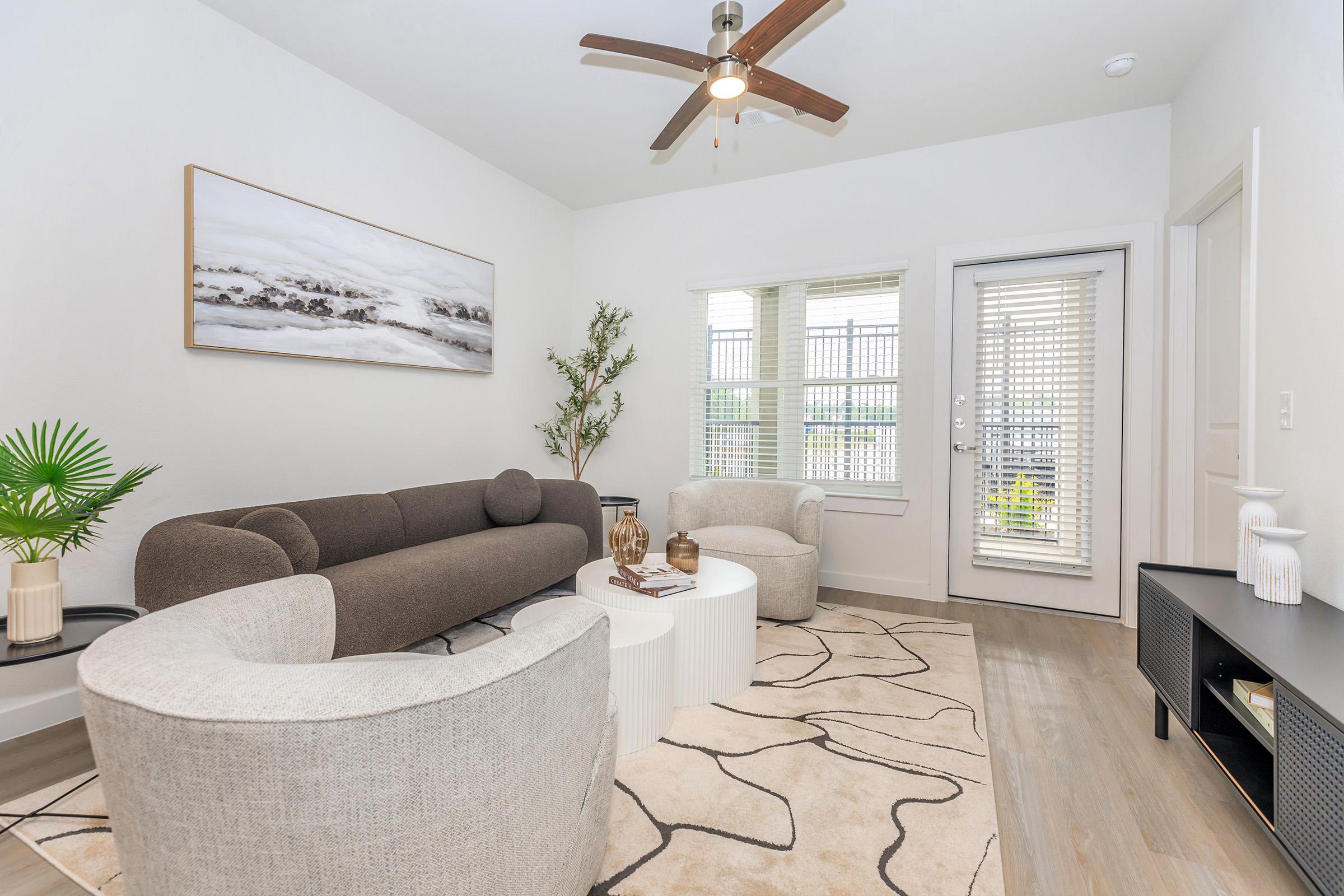 A modern living room featuring a gray and beige color scheme. A dark sectional sofa and two light, curved chairs surround a white coffee table. A decorative plant and vases add a touch of greenery. Natural light streams through a window with vertical blinds, and there is a door leading outside.