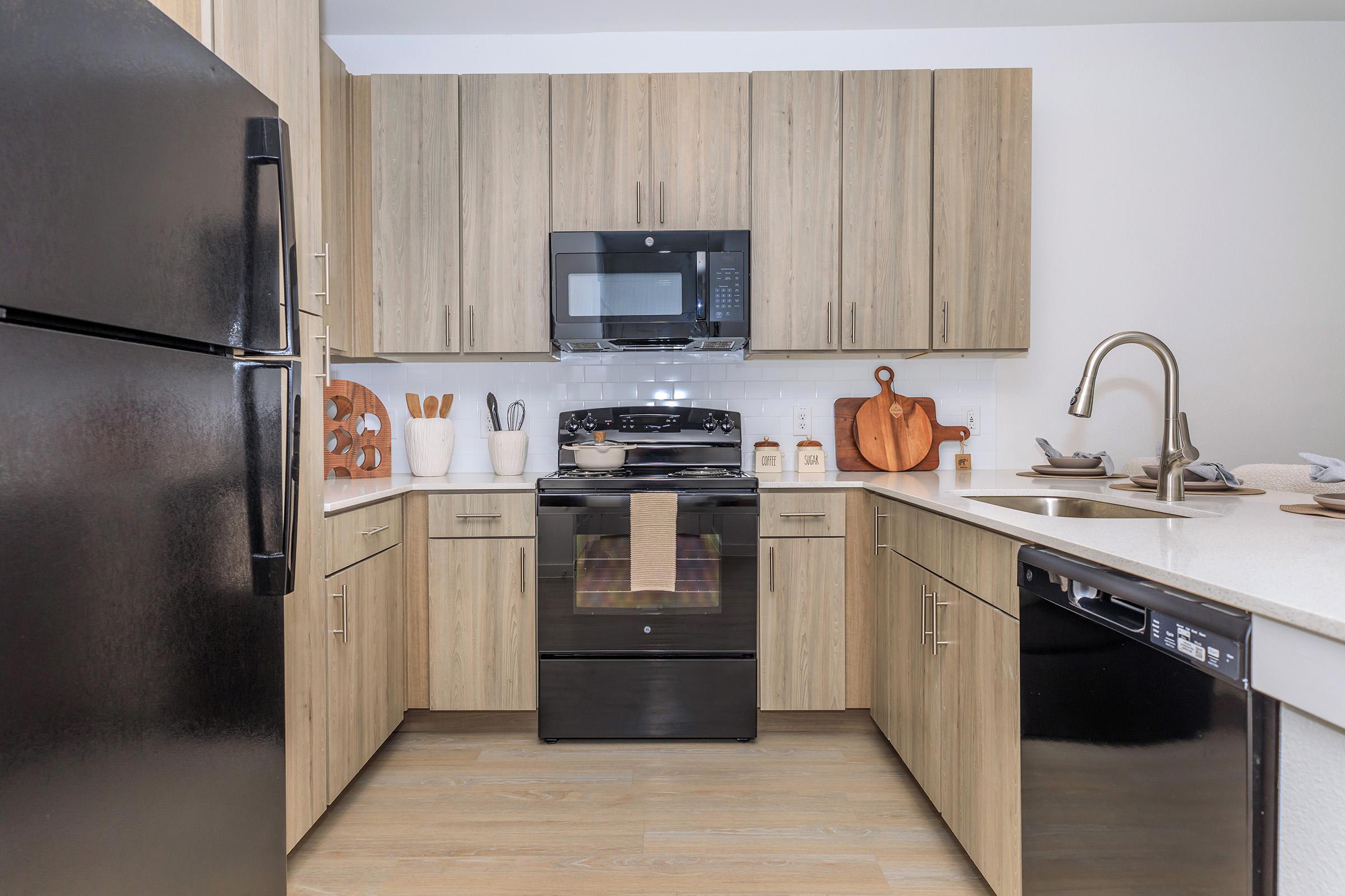 Modern kitchen featuring wooden cabinetry, black appliances including a refrigerator and an oven, a microwave above the stove, and a sink with a stylish faucet. Countertops are light-colored with decorative kitchenware, cutting boards, and utensils arranged neatly for a functional and inviting space.