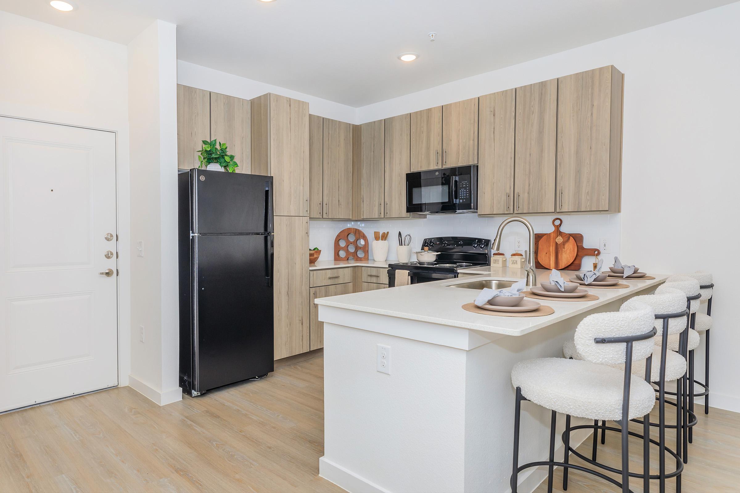 Modern kitchen featuring light wood cabinets, a black refrigerator, and a black microwave. A white island bar with four stools is positioned in front, set with plates and utensils. The decor includes a cutting board and some kitchen items on display, with a bright, airy feel from the natural light.