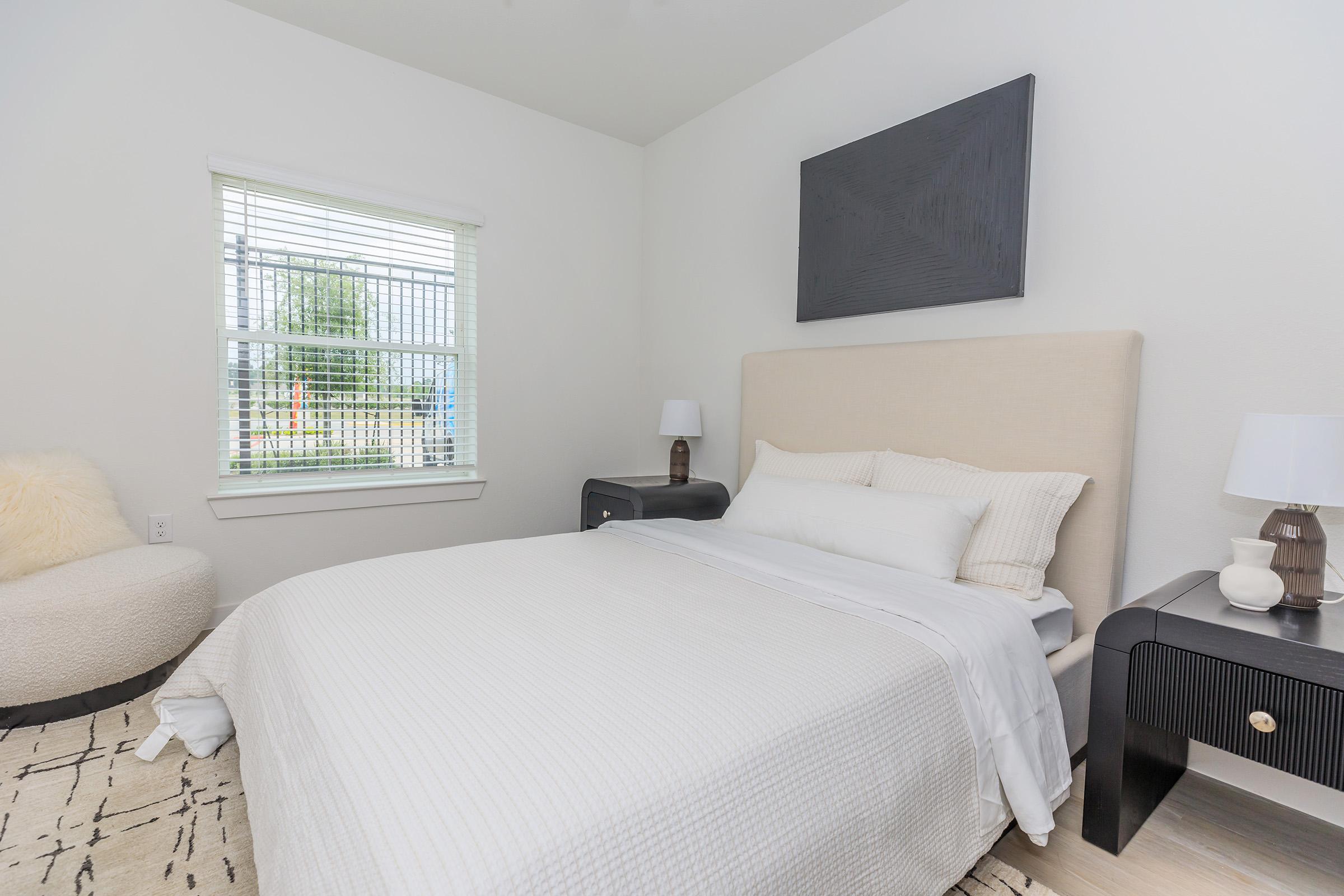 A cozy bedroom featuring a light-colored bed with white bedding, a beige upholstered headboard, and two bedside tables with lamps. There is a plush chair in one corner and a window with blinds letting in natural light, creating a bright and airy atmosphere. The walls are painted white, enhancing the spacious feel.