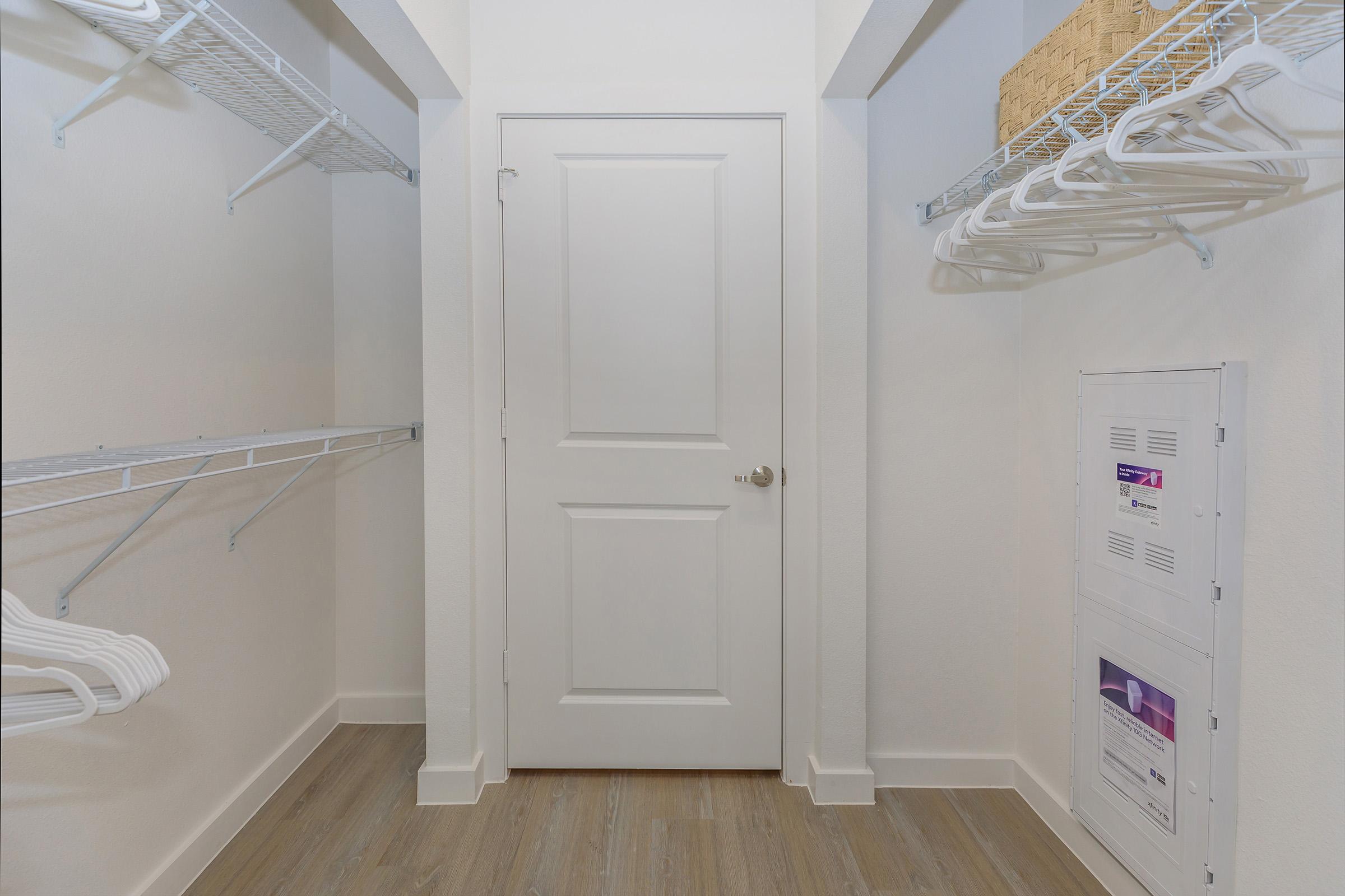 Empty closet space with white wire shelving and a closed white door. The flooring is light-colored wood, and there is a small basket on a shelf. There’s also a utility panel on the wall next to the door. Overall, the space is bright and minimalistic.