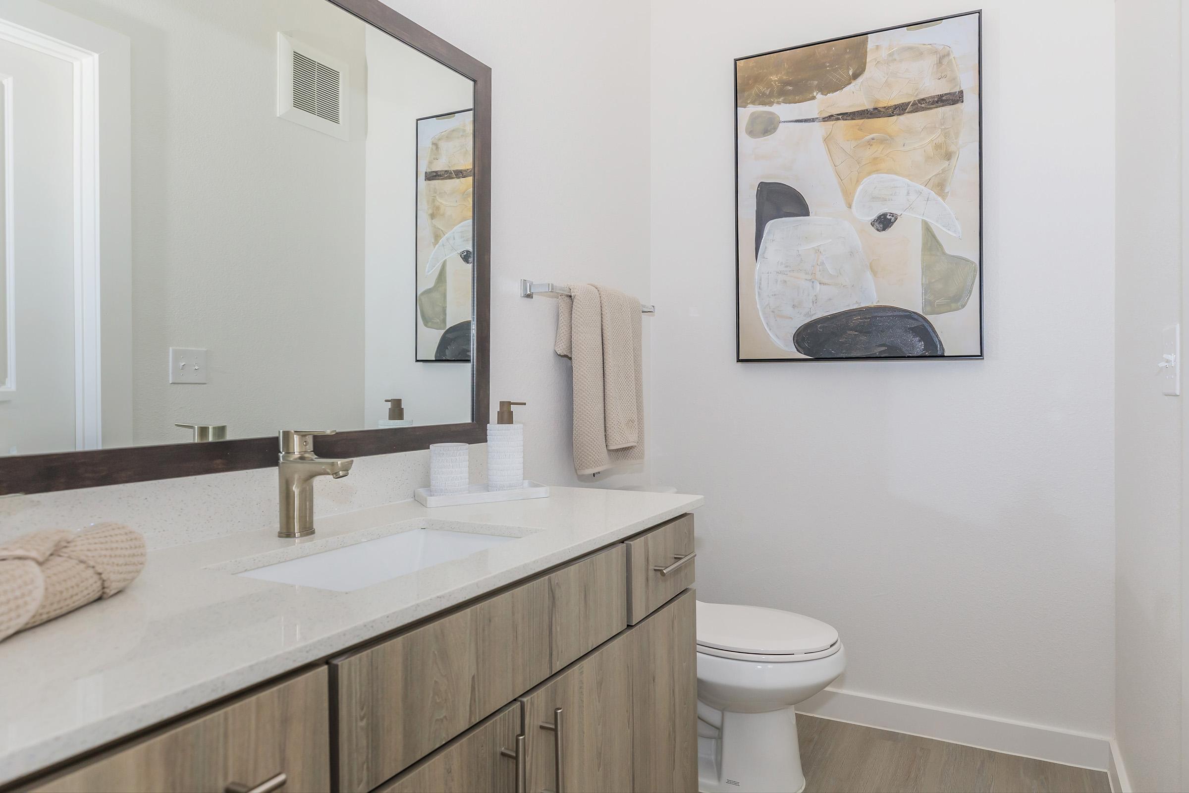 A modern bathroom featuring a wooden vanity with a sink, a large mirror, and a towel hanging on a rack. An abstract painting hangs on the wall above the vanity. There is a white toilet in the corner, and the walls are painted in a light color, creating a clean and contemporary look.