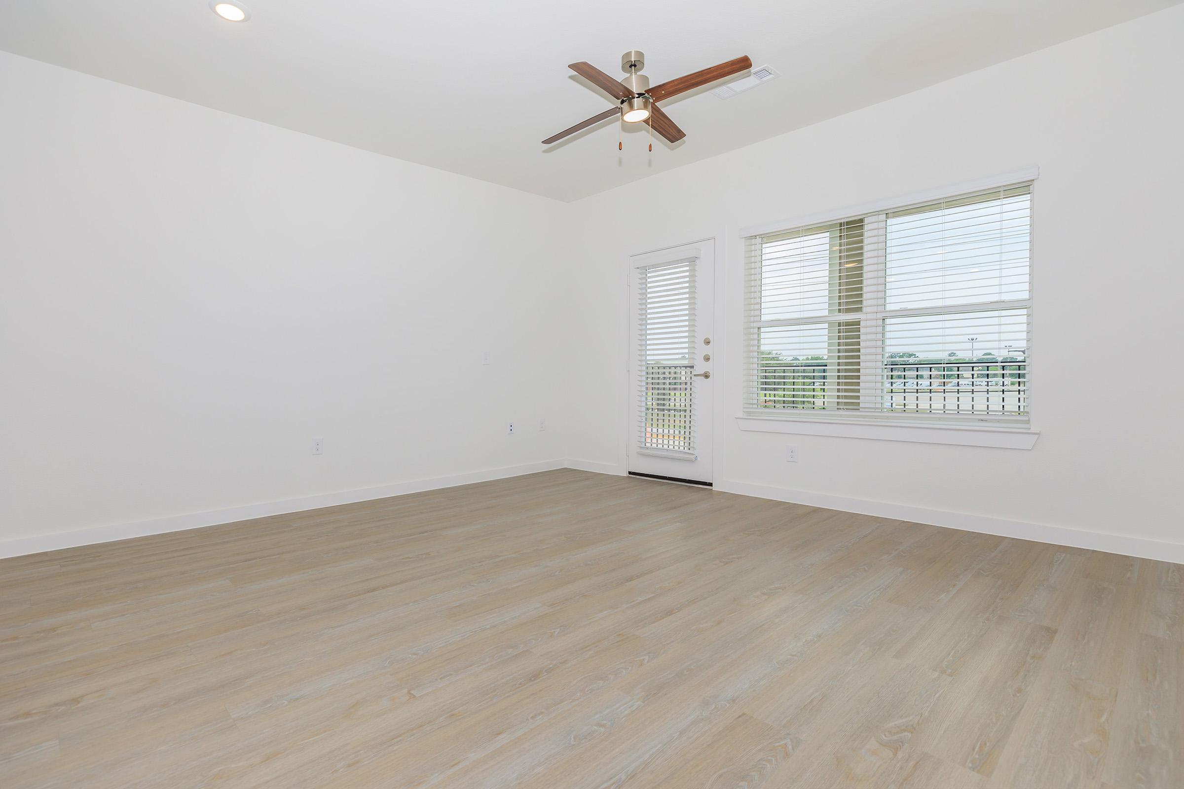 An empty room featuring light wood flooring, large windows with blinds, and a ceiling fan. There is a door leading outside, providing natural light and a view of the surroundings. The walls are painted white, creating a bright and airy atmosphere.