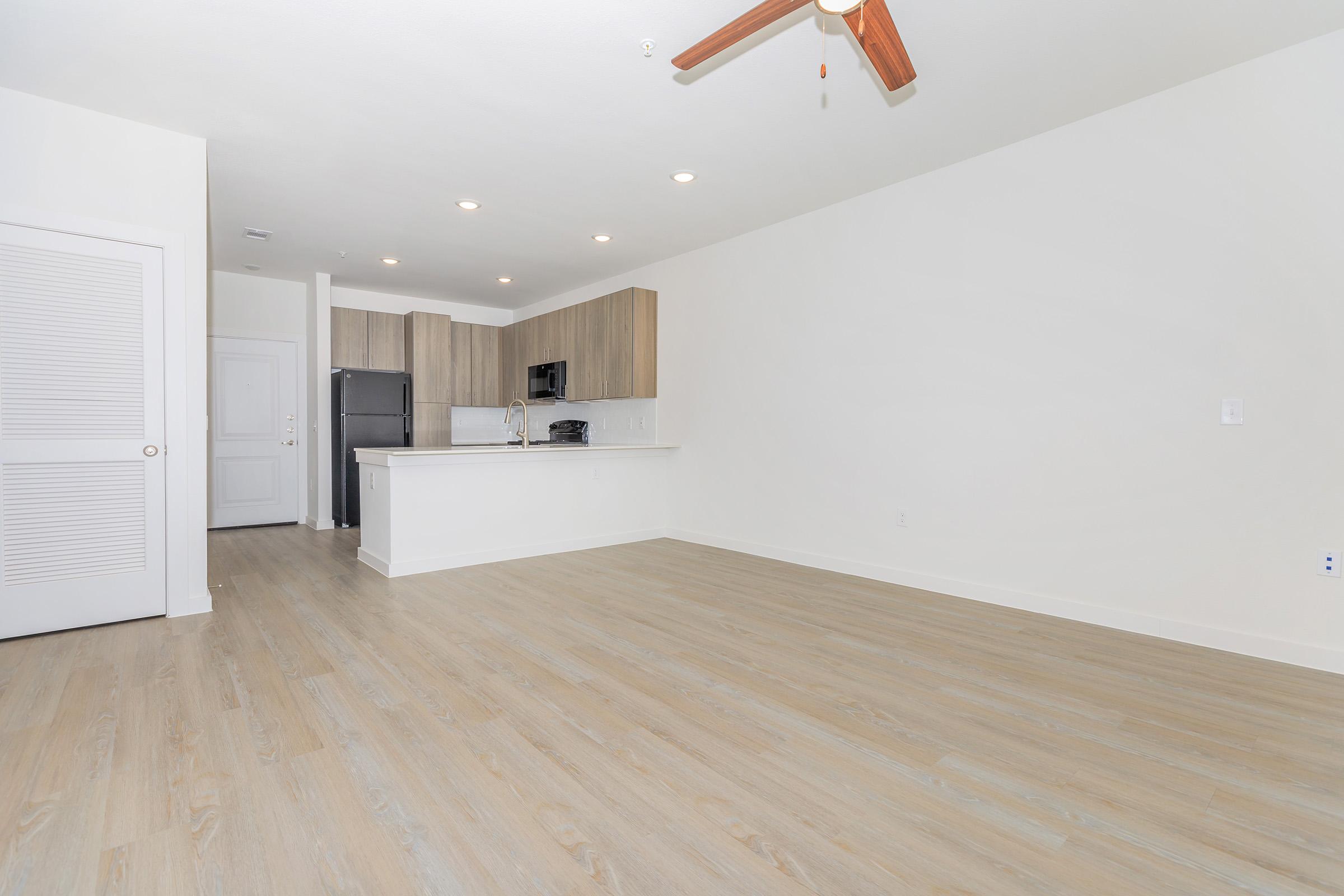 Spacious living area with light wood flooring, modern ceiling fan, and large windows. An open kitchen features wooden cabinets, stainless steel appliances, and a counter space for seating. The design is minimalistic with a bright, inviting atmosphere.