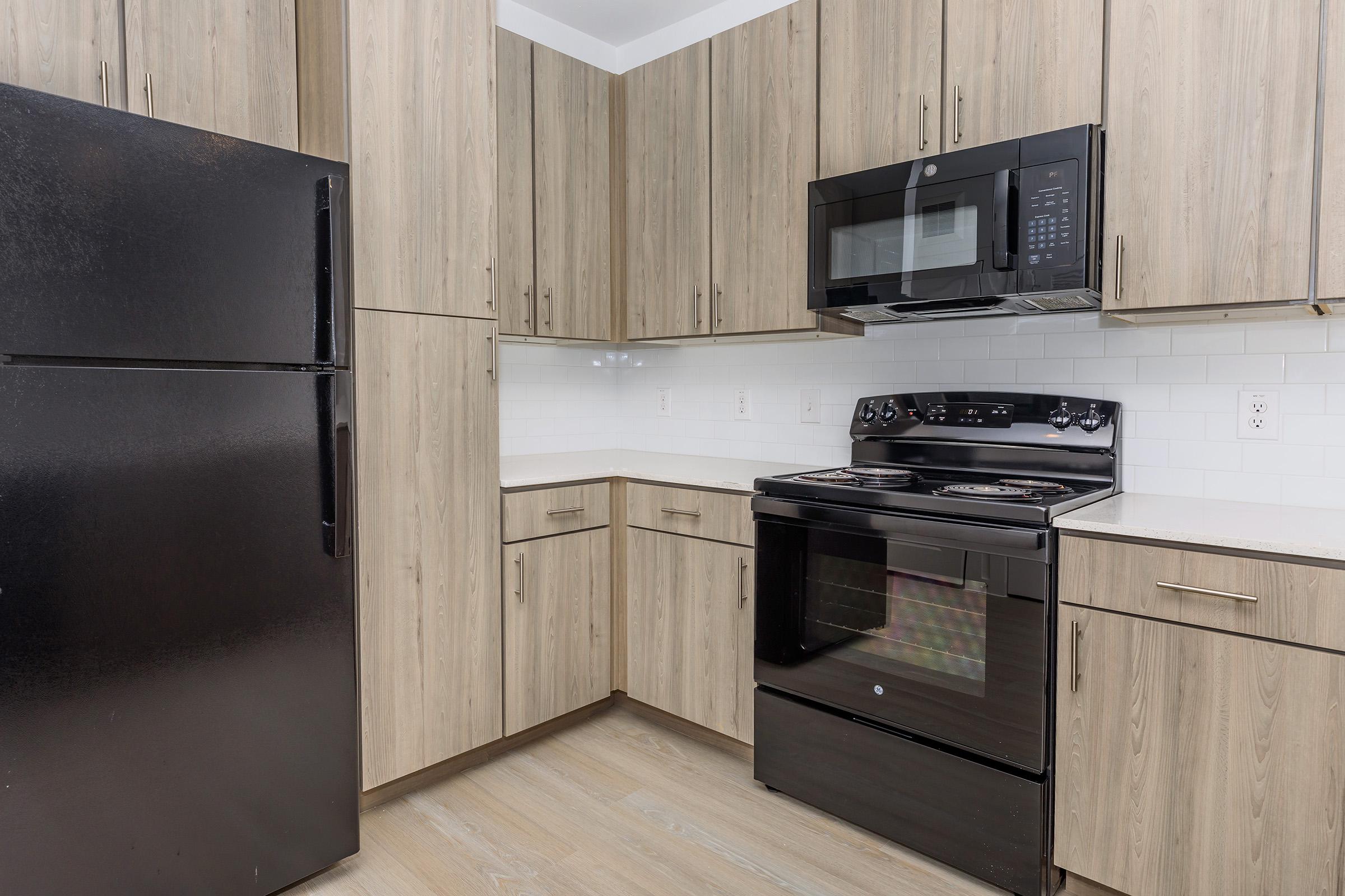 Modern kitchen featuring wooden cabinets, a black refrigerator, and a black stove with an oven. Above the stove, there is a black microwave built into the cabinetry. The countertop is light-colored, and the backsplash consists of white tiles. The flooring is a light, neutral tone.