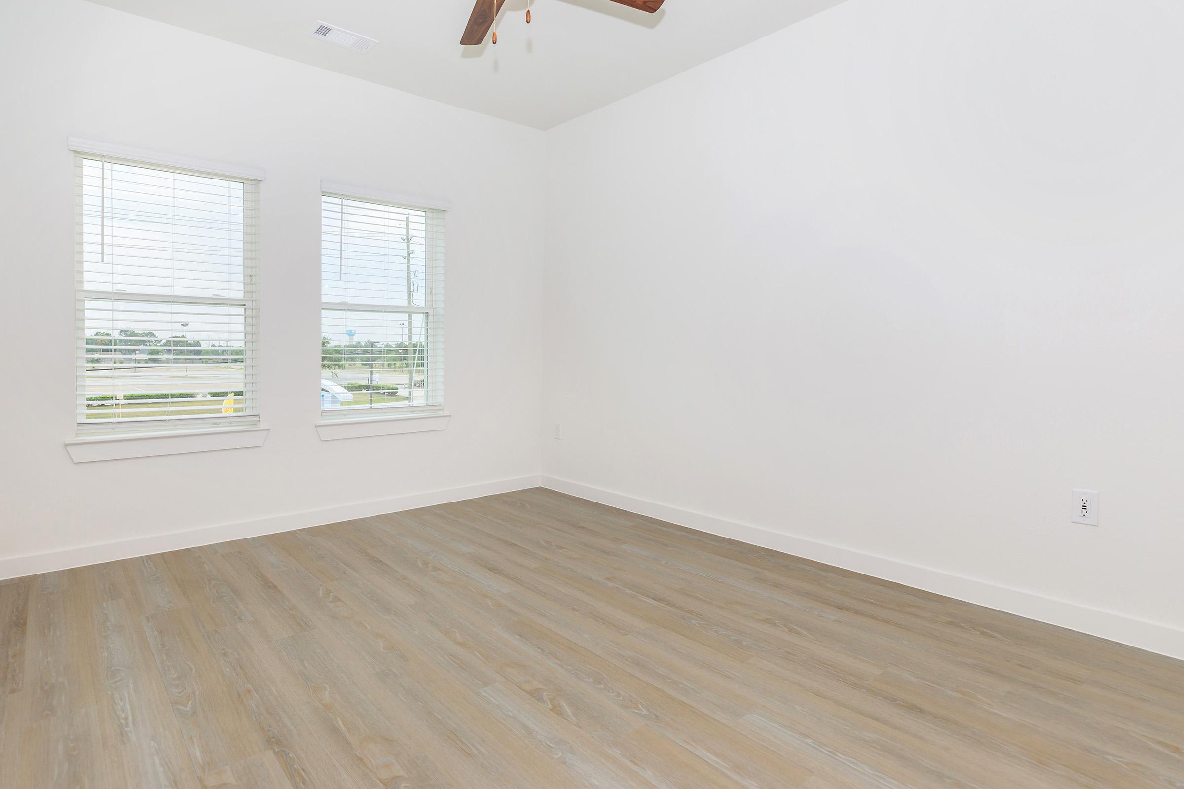 A bright and empty room featuring light-colored walls, two windows with blinds, and a light wooden floor. The room has a ceiling fan but no furniture, creating a spacious and minimalistic look. Natural light fills the space, giving it a fresh and inviting atmosphere.