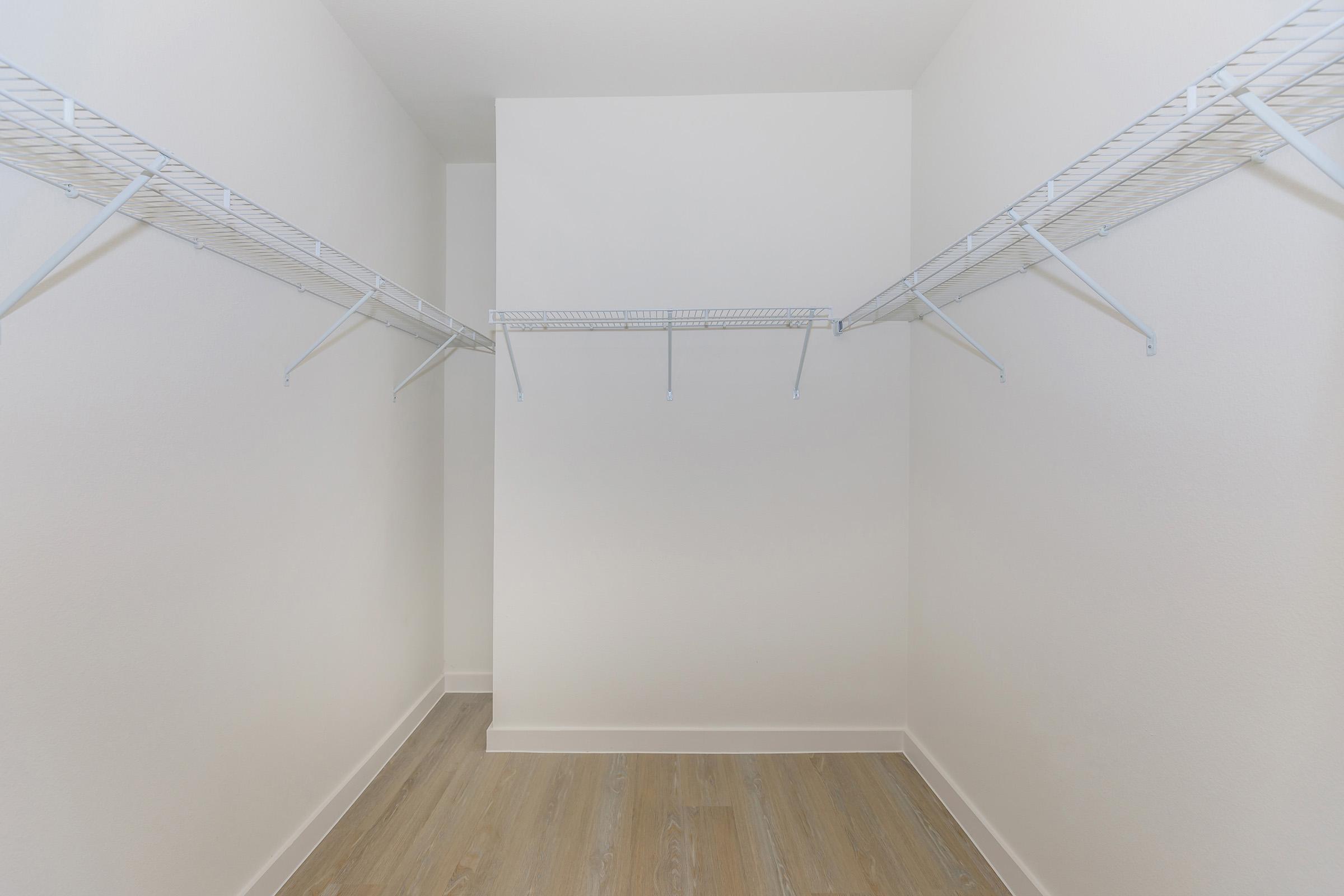 Empty walk-in closet with white wire shelving on two sides, light gray walls, and a wooden floor. The space is bright and minimalistic, providing ample room for storage.