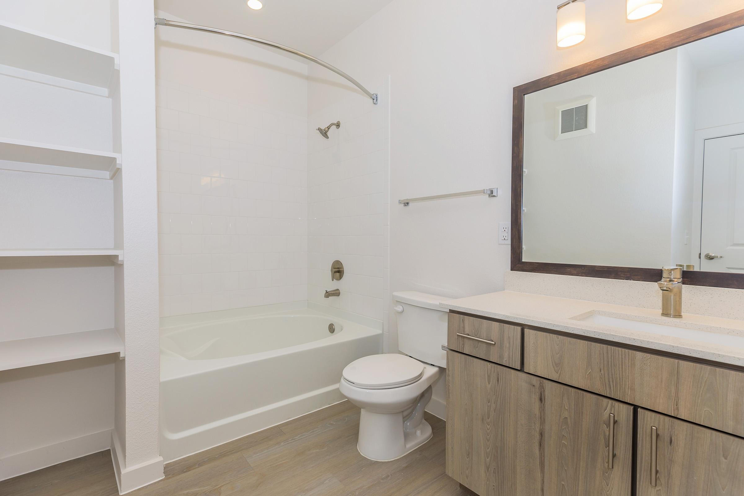 Modern bathroom featuring a bathtub with shower, toilet, and a vanity with a sink. The walls are white, and there is a large mirror above the vanity. Light wood cabinets provide storage, and there are open shelves for additional space. The flooring is light wood, creating a clean and contemporary look.