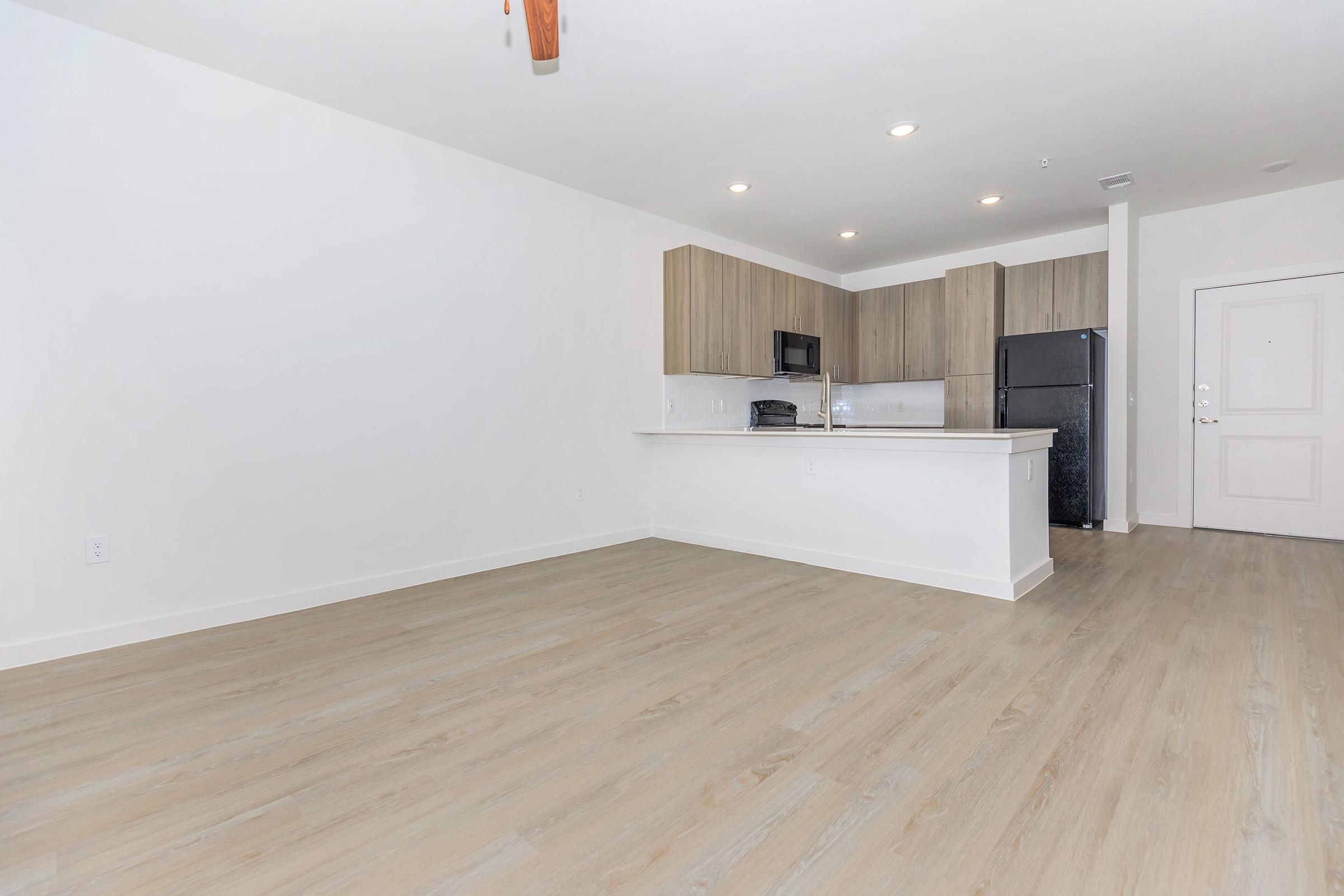 A modern, spacious kitchen and living area featuring light wood floors, white walls, and a minimalist design. The kitchen includes wooden cabinets, stainless steel appliances, and a countertop bar. Natural light from an unseen window creates a bright atmosphere in the room.