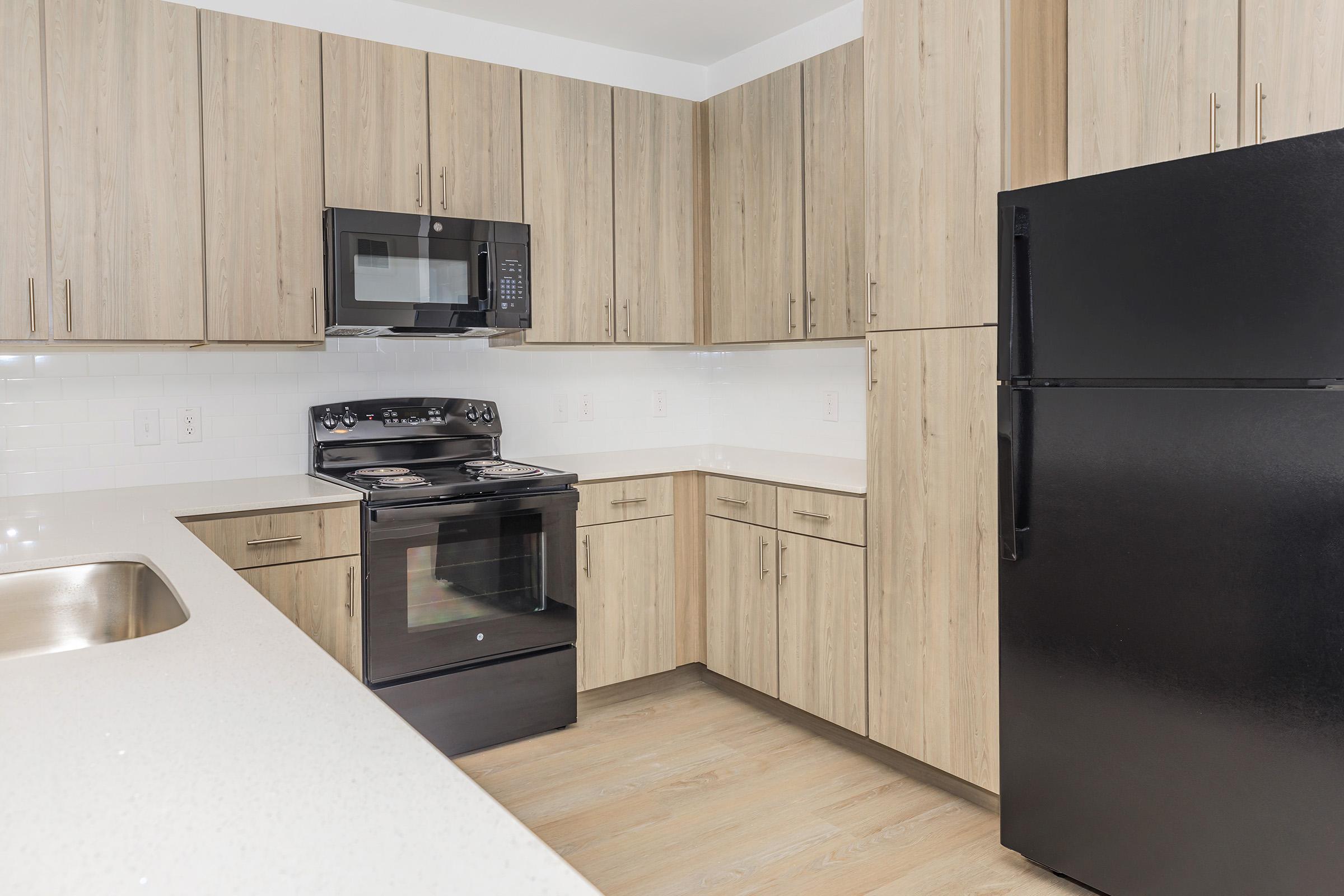 A modern kitchen featuring light wood cabinetry, a black refrigerator, a black microwave oven, and a stainless steel gas stove. The countertops are white with a subtle texture, and the space has a clean, minimalist design with ample storage and sleek finishes.