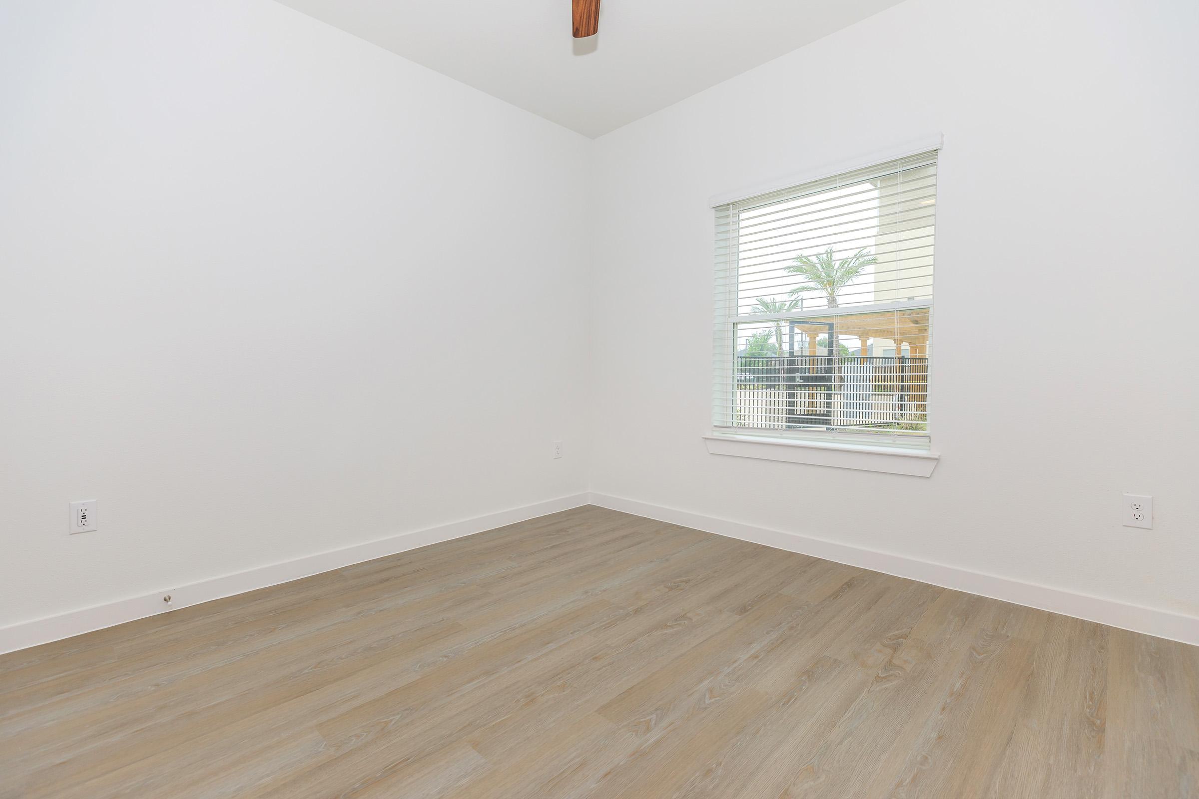 Empty room with light-colored walls and a single window featuring blinds. The flooring is a light wood tone, and there's a ceiling fan. The room has no furniture, creating a spacious and minimalistic appearance.