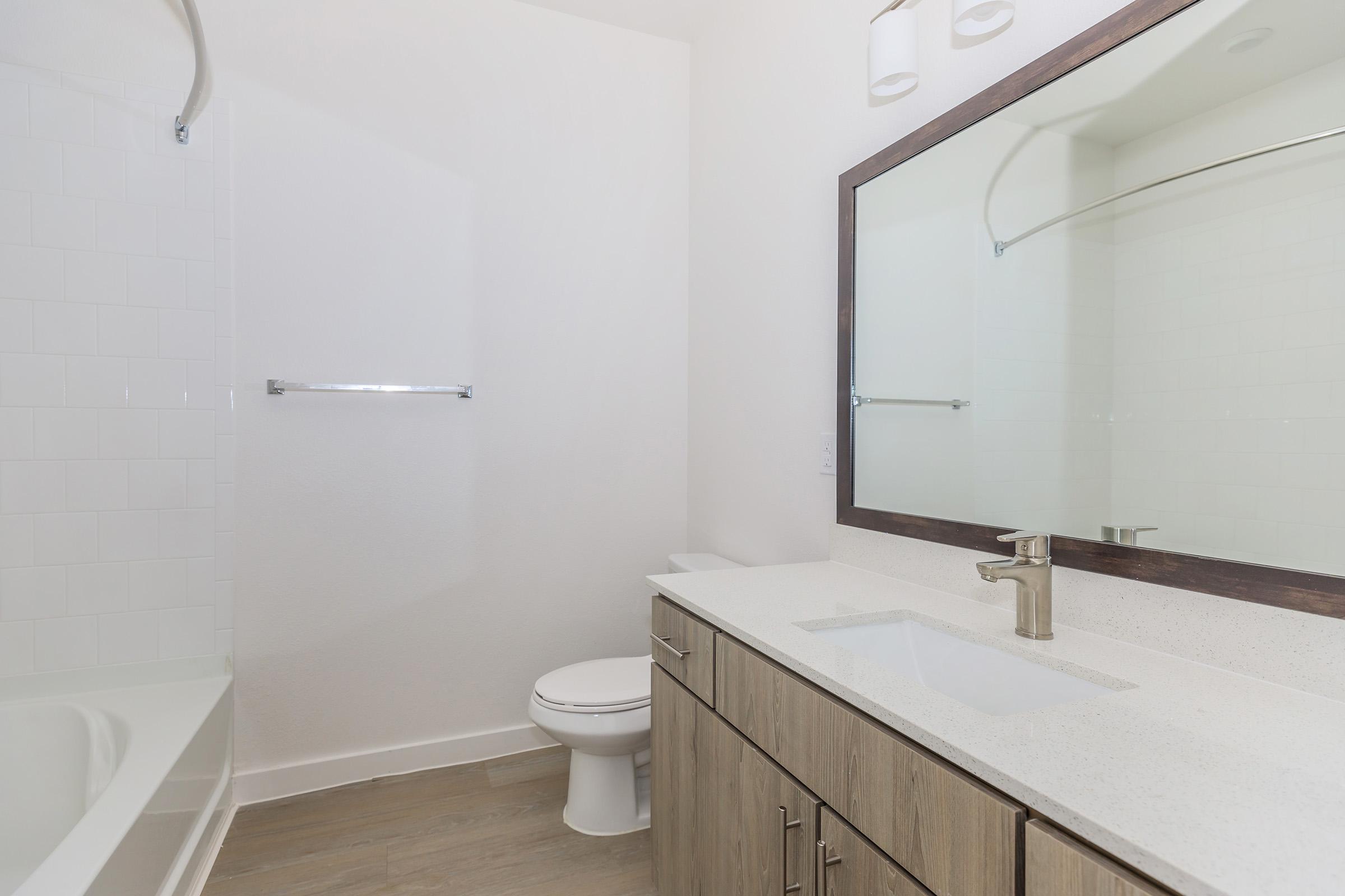 A modern bathroom featuring a bathtub, toilet, and a vanity with a sink. The vanity has light wooden cabinets and a large mirror above it. The walls are white, and the floor is light-colored. Natural light brightens the space, creating a clean and inviting atmosphere.