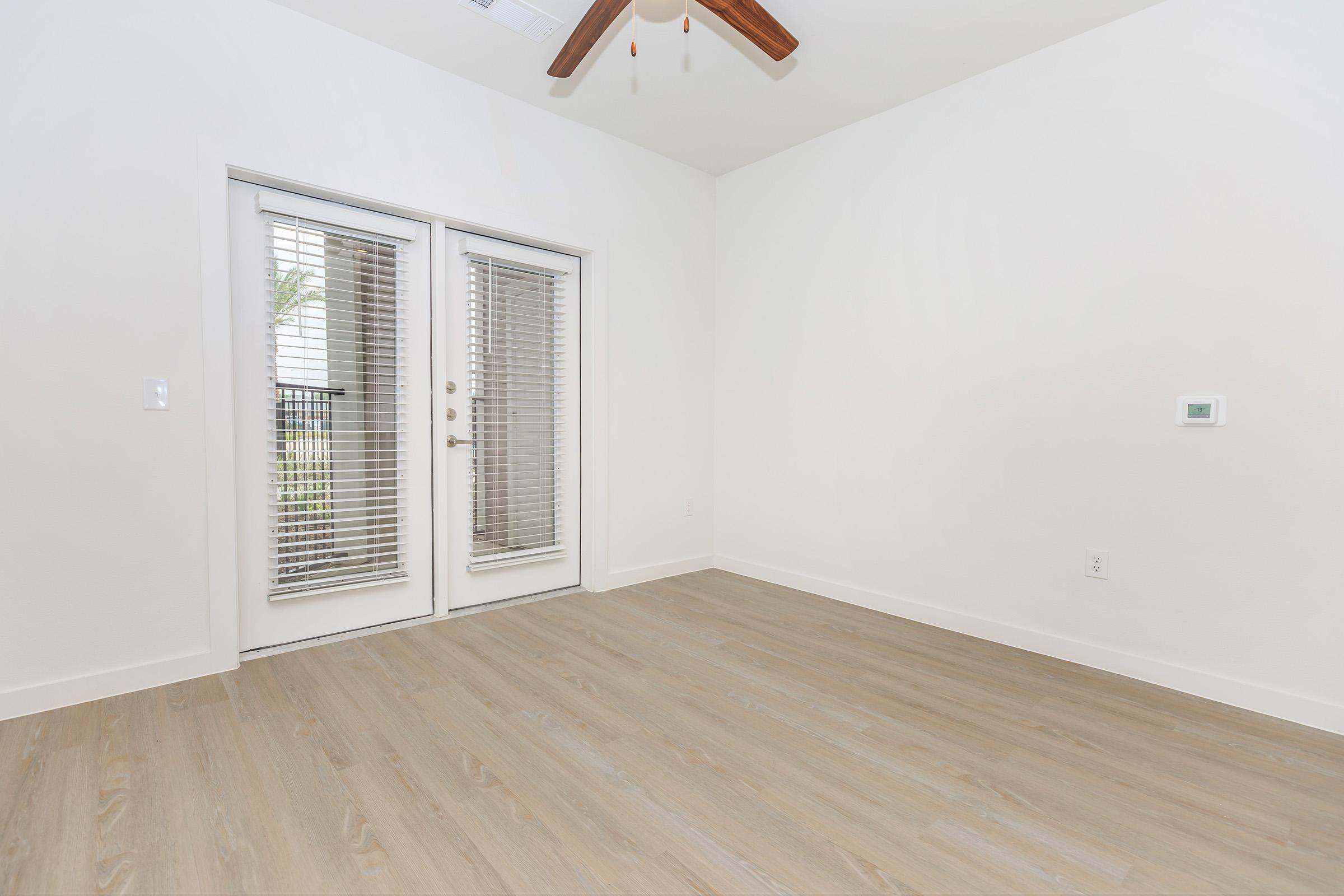 A spacious, empty room with light-colored walls and wooden flooring. There are double glass doors featuring blinds that lead to a balcony. A ceiling fan is visible above, and a small thermostat is mounted on the wall to the right. The room has natural light coming in through the doors.