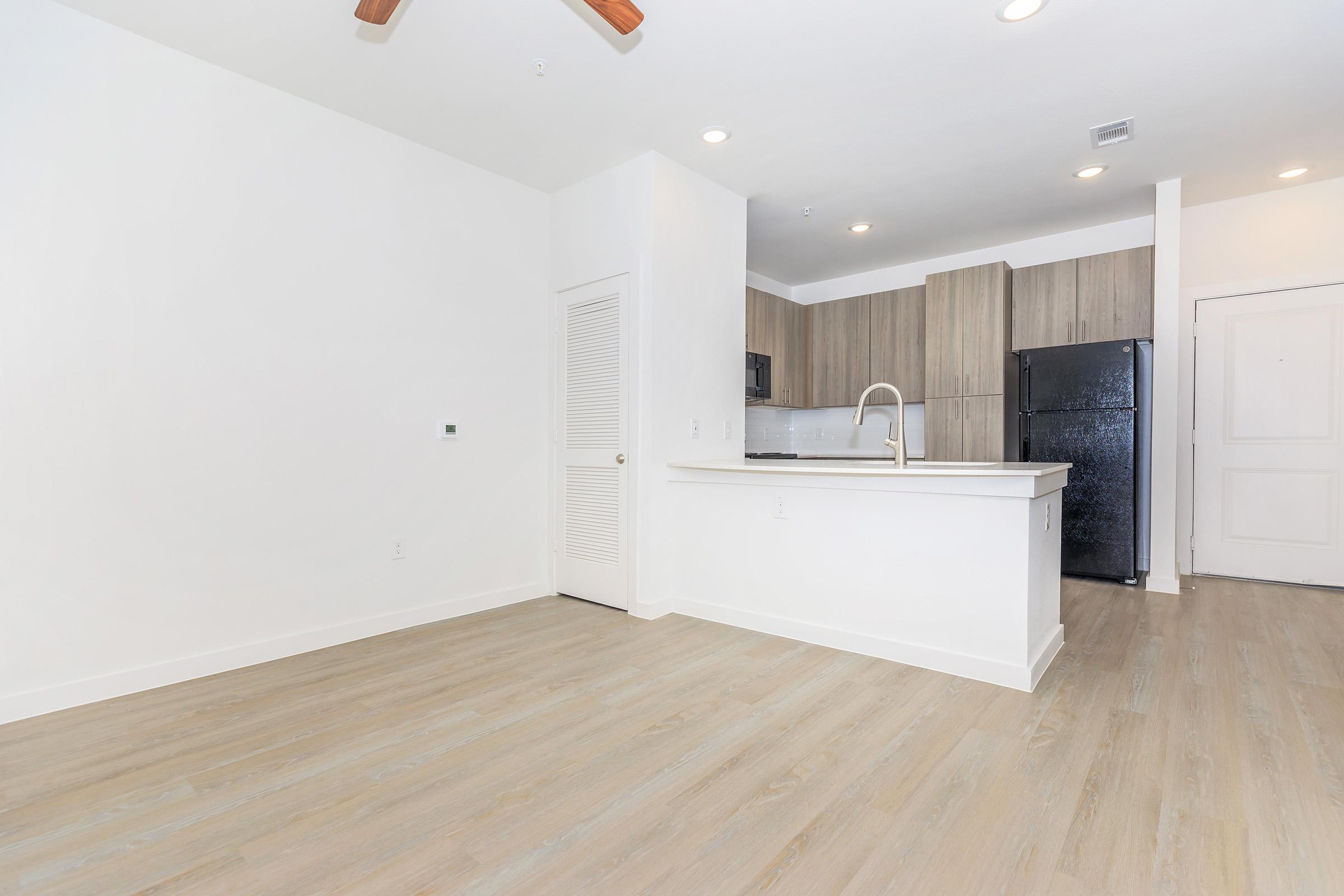 A well-lit, modern apartment interior featuring a white wall, light wood flooring, and a small kitchen area with dark appliances. The space includes a ceiling fan and an open layout, with a closed door to the left and a doorway leading out of view.