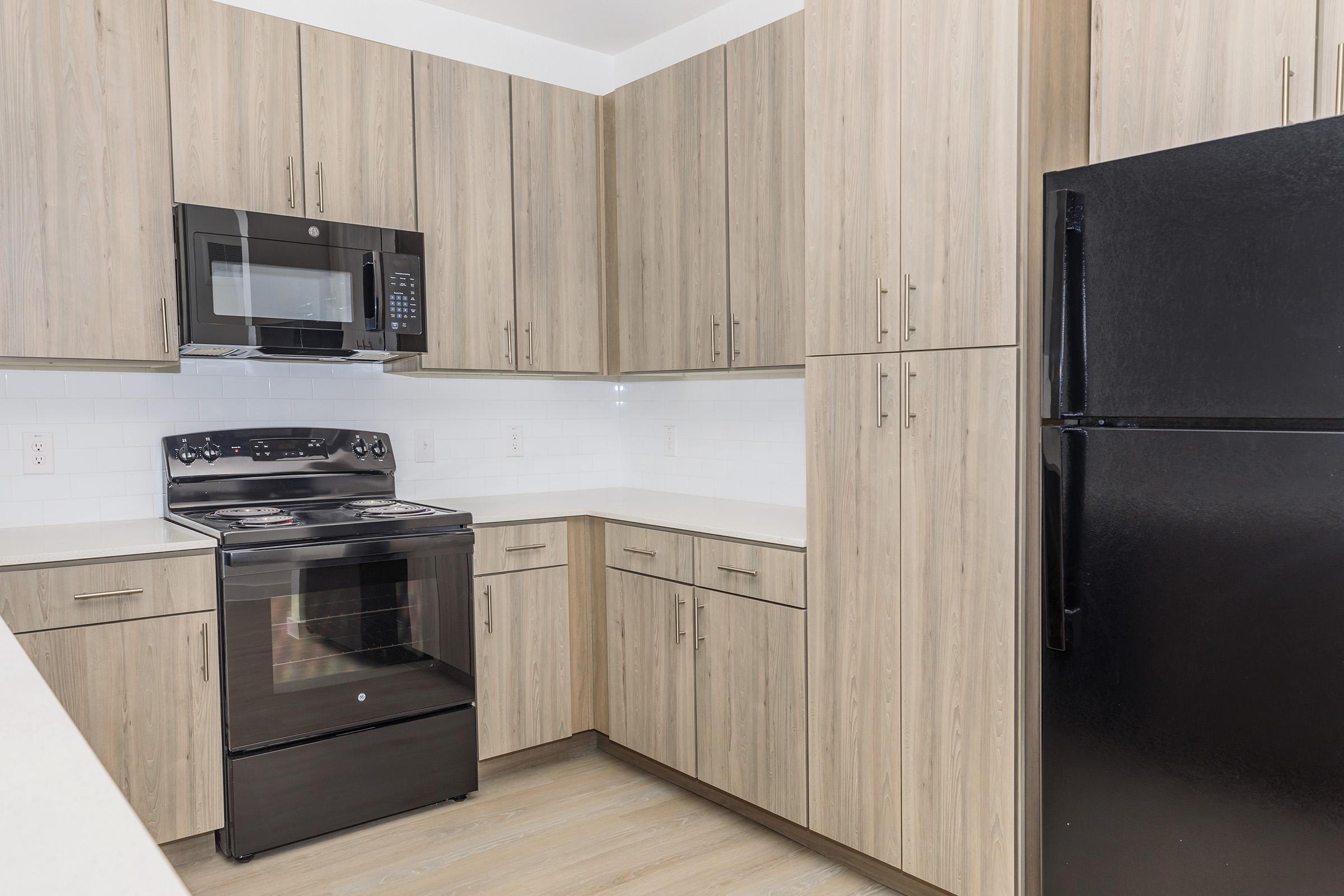 Modern kitchen with wooden cabinets, black appliances including a stove and microwave above, and a refrigerator. The countertop is light-colored, and the backsplash features white tiles. The flooring is a light wood tone, creating a contemporary and spacious look.