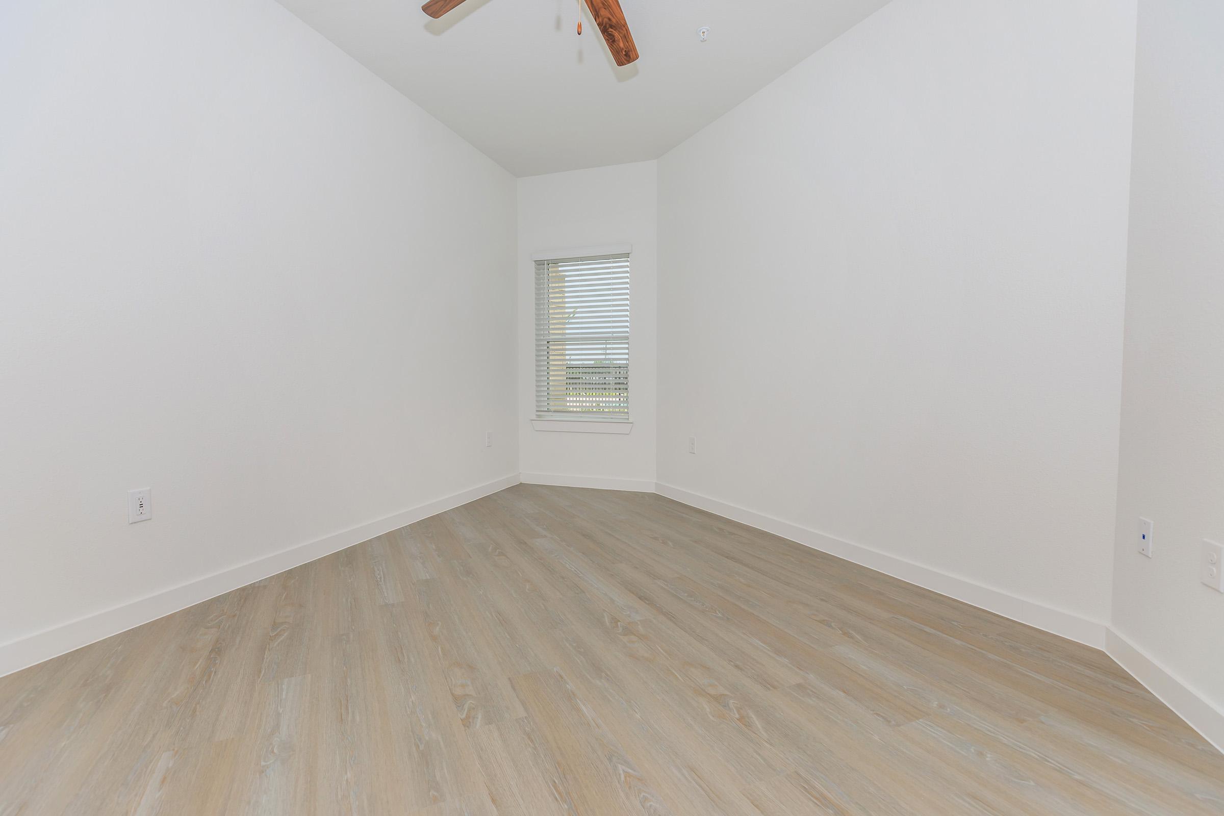 A brightly lit, unfurnished room with white walls and light wood flooring. A single window with blinds allows natural light to enter, and the ceiling features a ceiling fan. The room has an open feeling, suitable for various furniture arrangements.