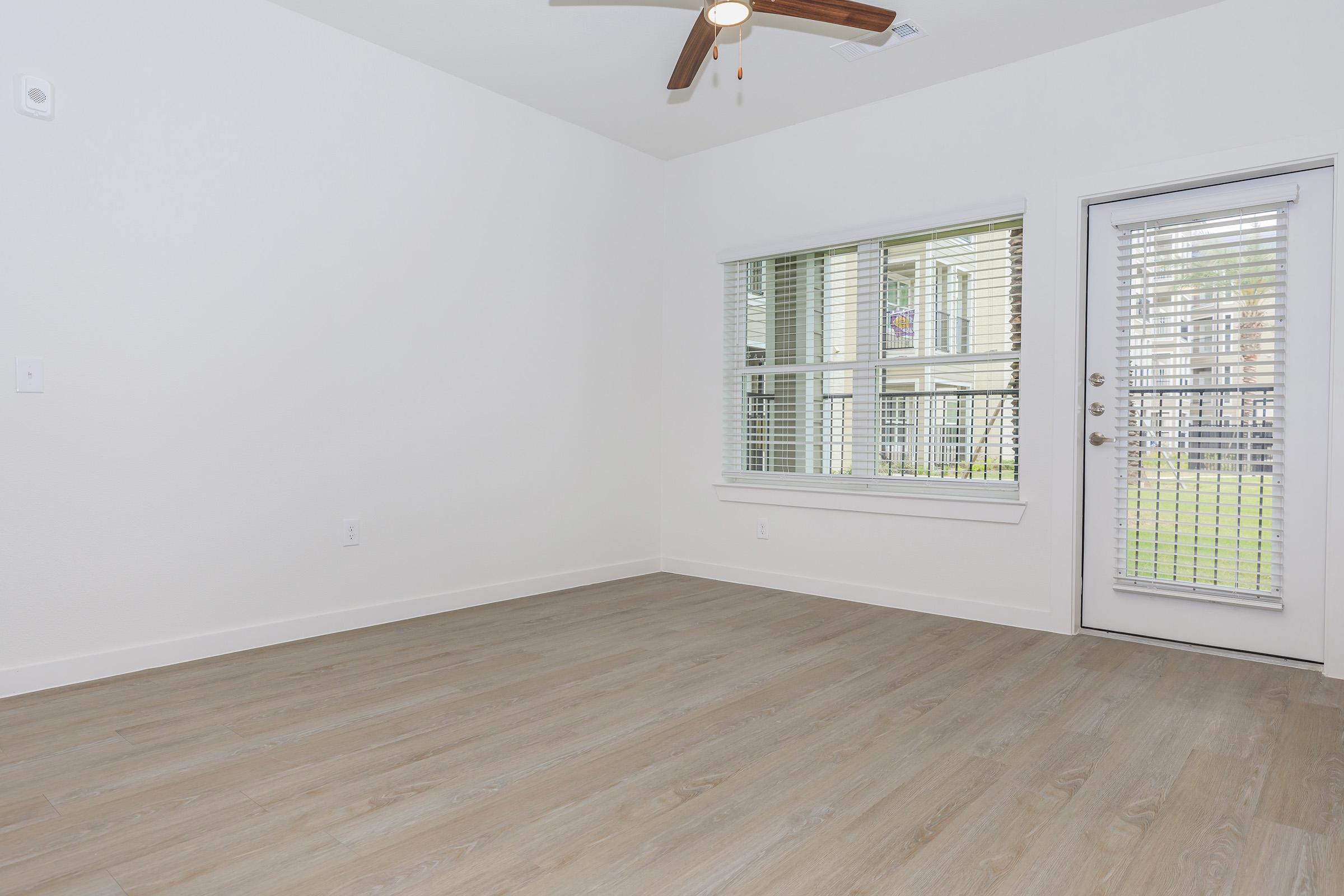 A bright, empty room featuring light wood flooring, a ceiling fan, and a large window with blinds. The window looks out to an outdoor area. A door with a window panel leads to the outside, offering ample natural light, while the walls are painted in a clean, neutral tone.