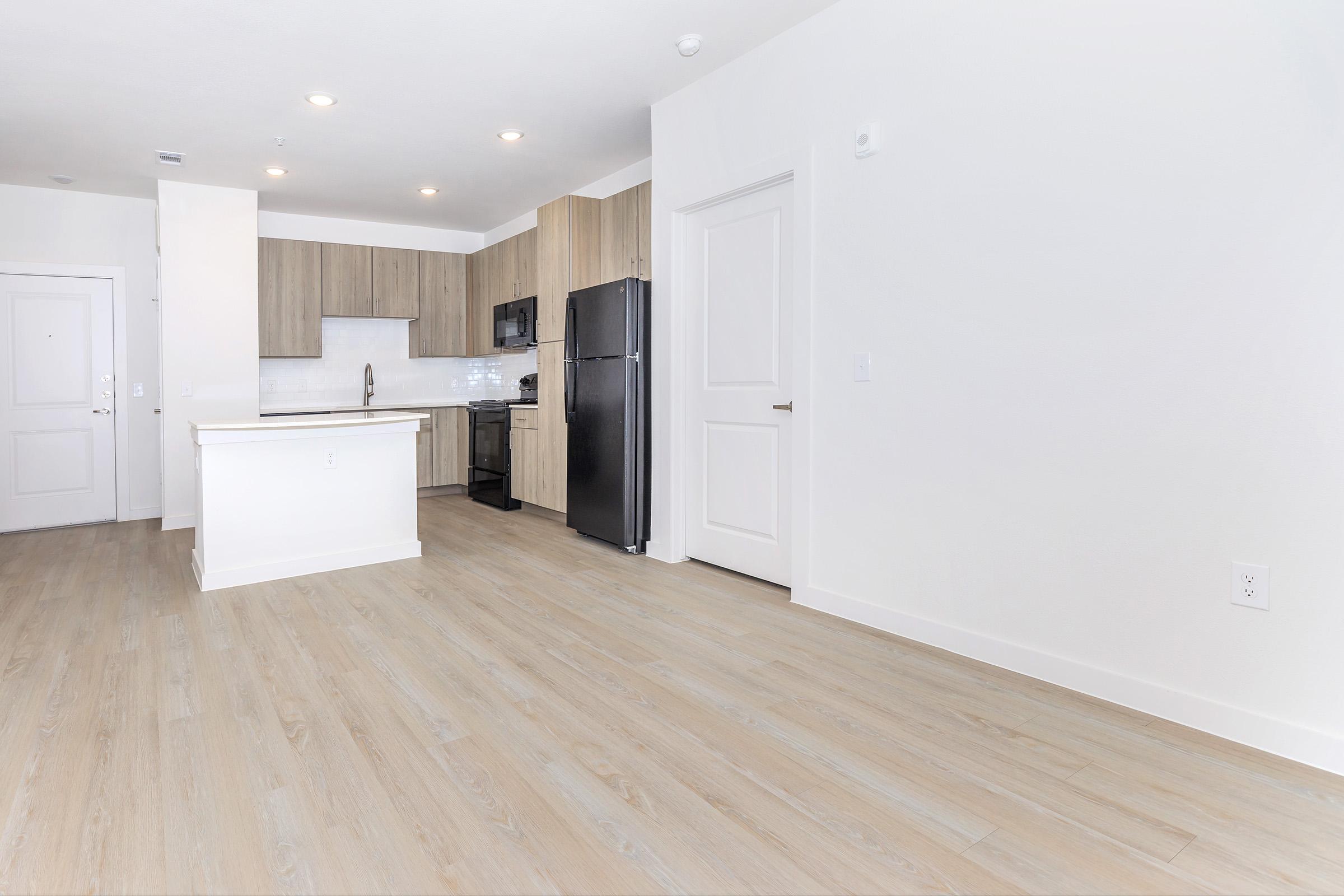Interior view of a modern kitchen and living area featuring light-colored wooden flooring, a spacious layout, a kitchen island with bar seating, and appliances including a black refrigerator and oven. The walls are painted in soft neutral tones, and there is a doorway leading to another room.