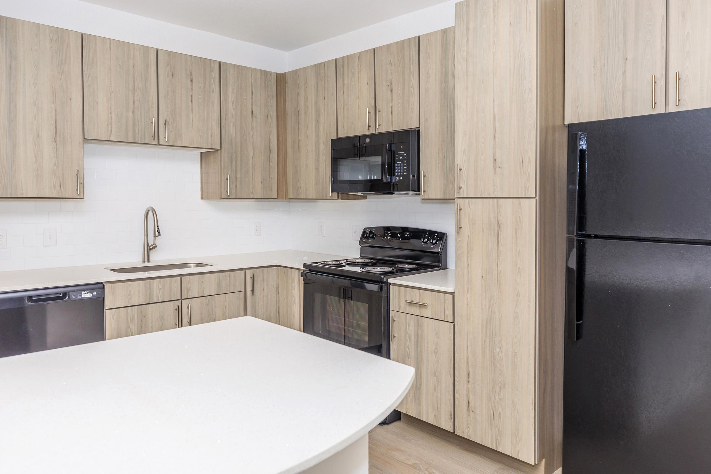 A modern kitchen featuring light wood cabinetry, a white countertop, and stainless steel appliances, including a microwave, oven, and refrigerator. The layout includes a sink and an island, with sleek, contemporary lines and a bright, clean aesthetic.