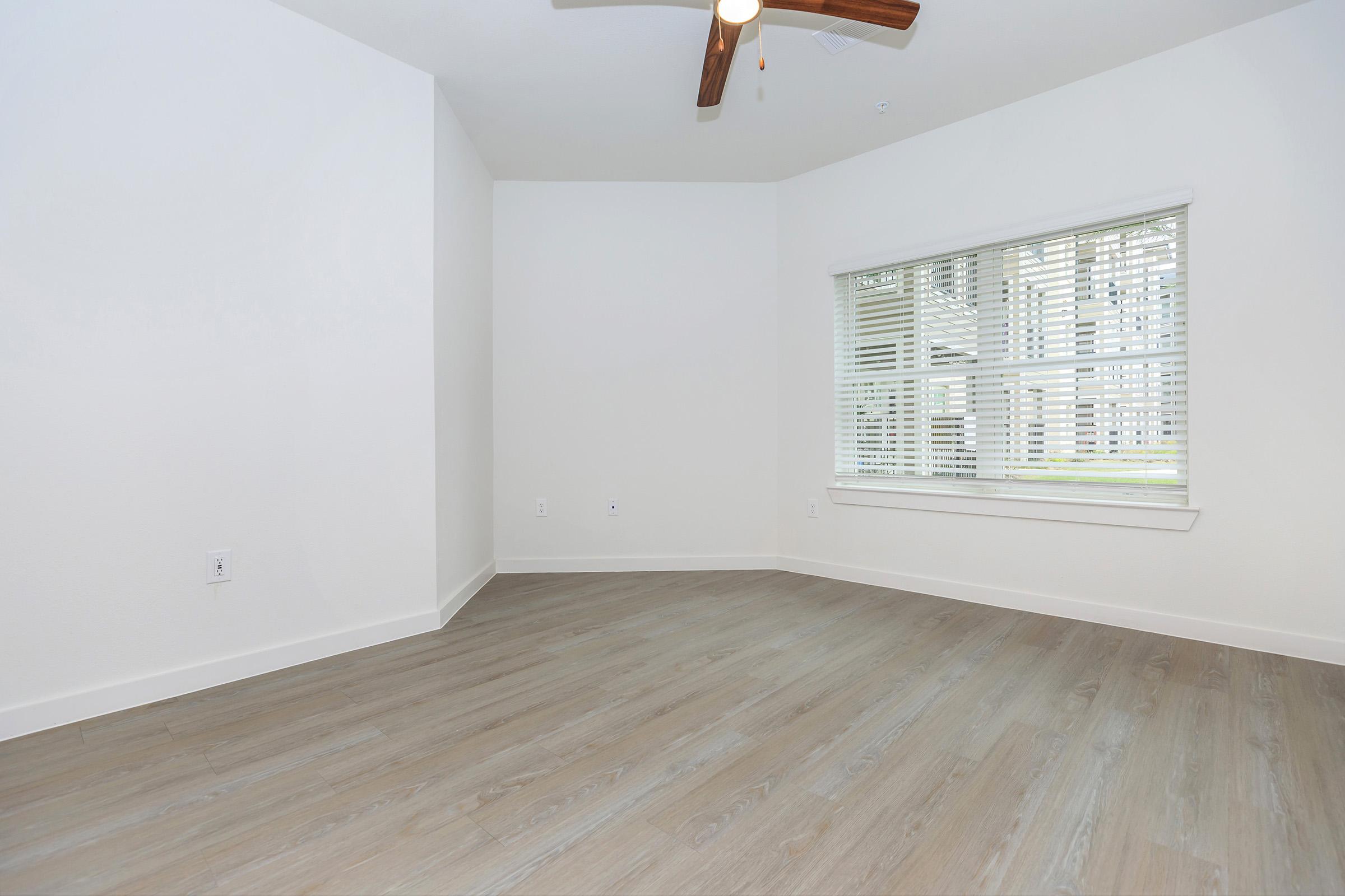 Empty room with light-colored walls and a wooden floor. A window with blinds allows natural light to enter, and a ceiling fan is visible. The space is clean and uncluttered, suggesting a fresh and modern aesthetic.