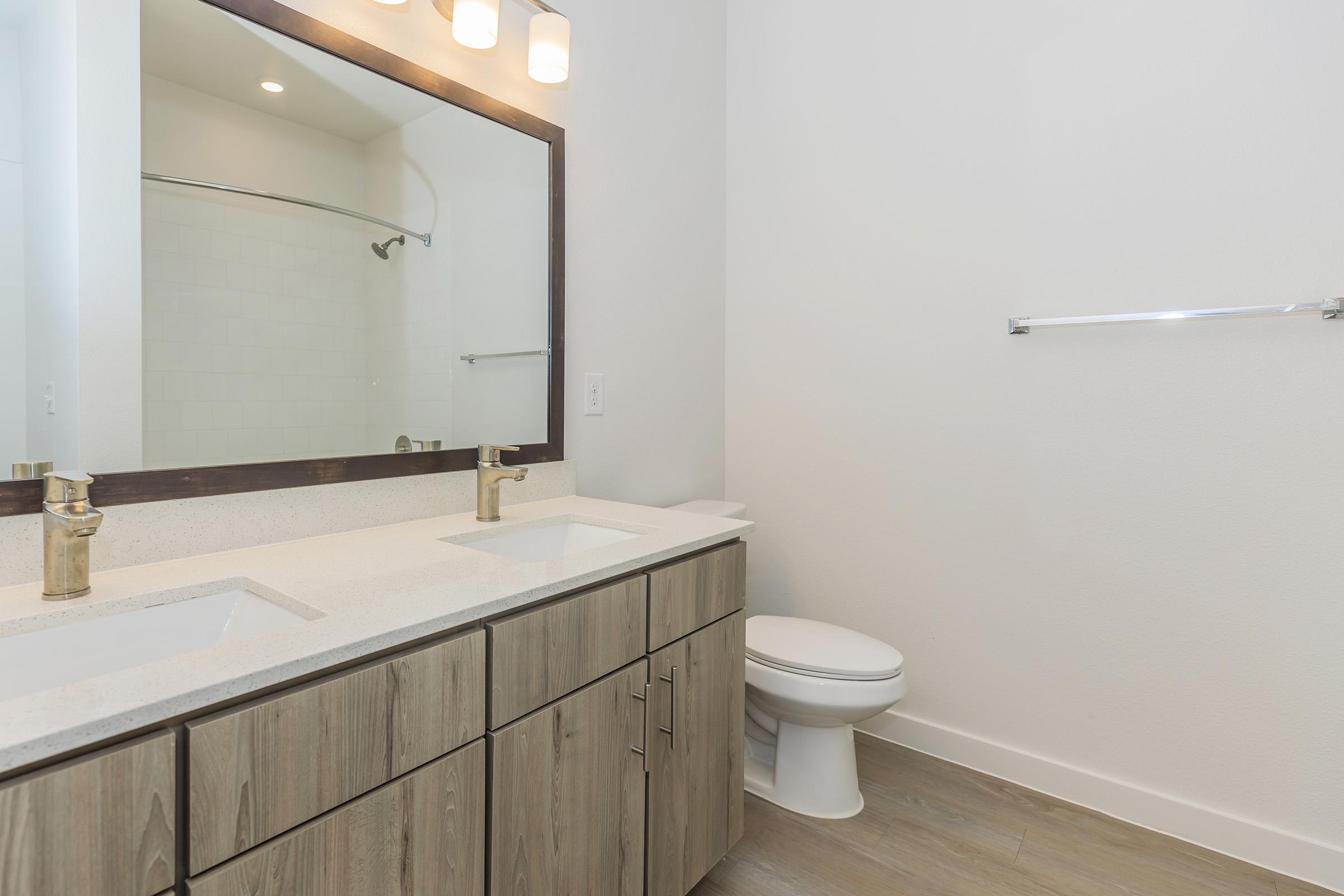 A modern bathroom featuring a double vanity with gold faucets, a large mirror above, and a toilet. The walls are painted white, and there is a light wooden floor. A glass-enclosed shower is visible in the background, adding to the contemporary design.