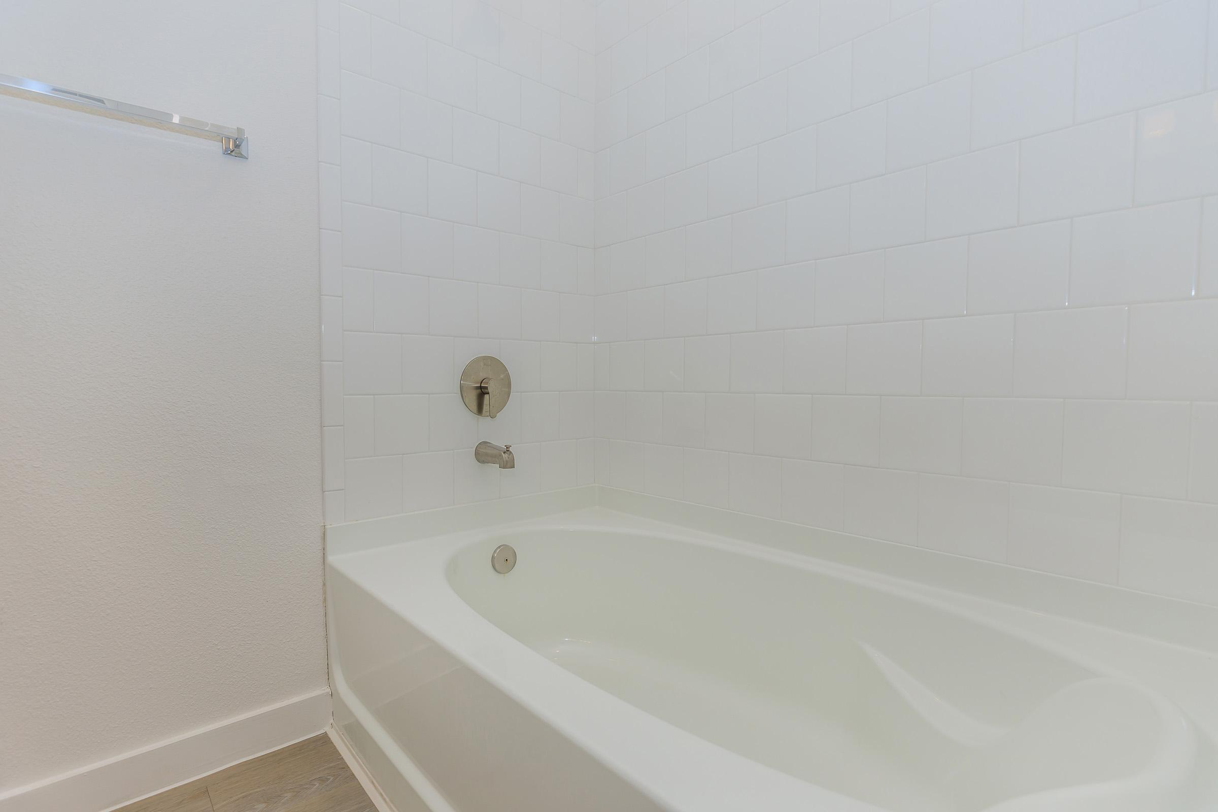A clean, modern bathroom featuring a white bathtub with a sleek silver faucet and wall-mounted showerhead. The walls are tiled with white tiles, and there is a shiny towel rack on the left side. The overall aesthetic is bright and minimalistic.