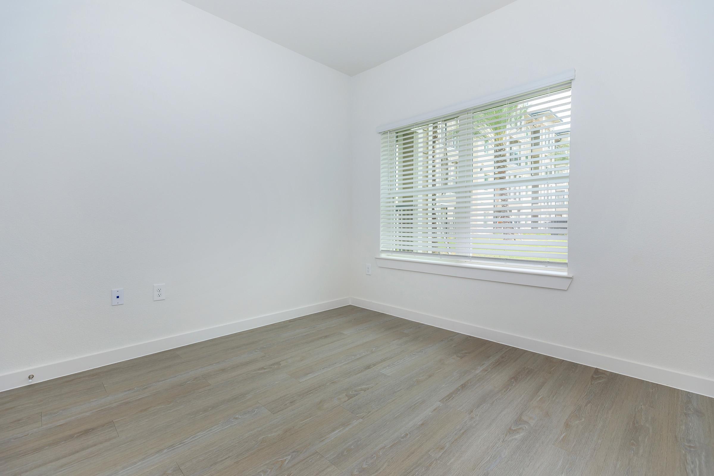 A minimally decorated room with light-colored walls and wooden flooring. There is a window with white blinds allowing natural light to enter, but no furniture or decorations are present, creating an empty and spacious feel.