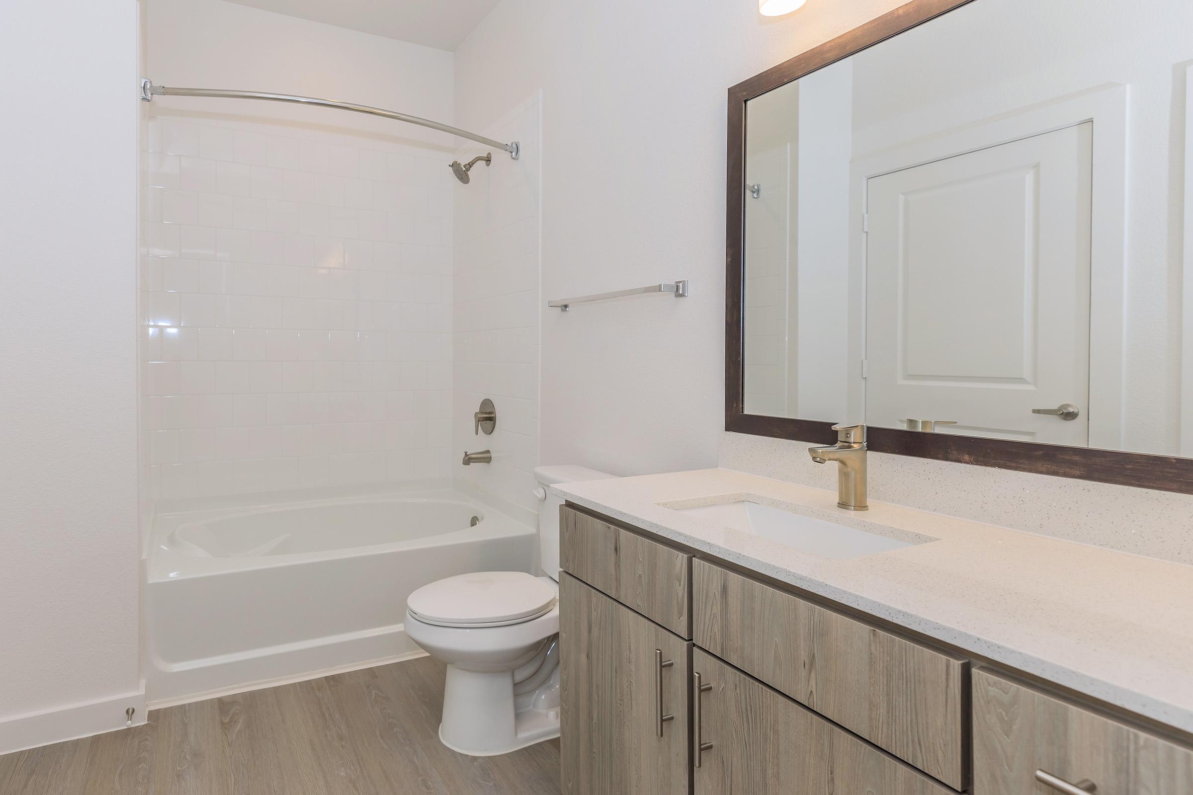 A modern bathroom featuring a bathtub with a shower, a toilet, and a double-sink vanity with wooden cabinetry. The space has neutral-colored walls and a large mirror above the sinks, complemented by warm lighting and a clean, contemporary design.
