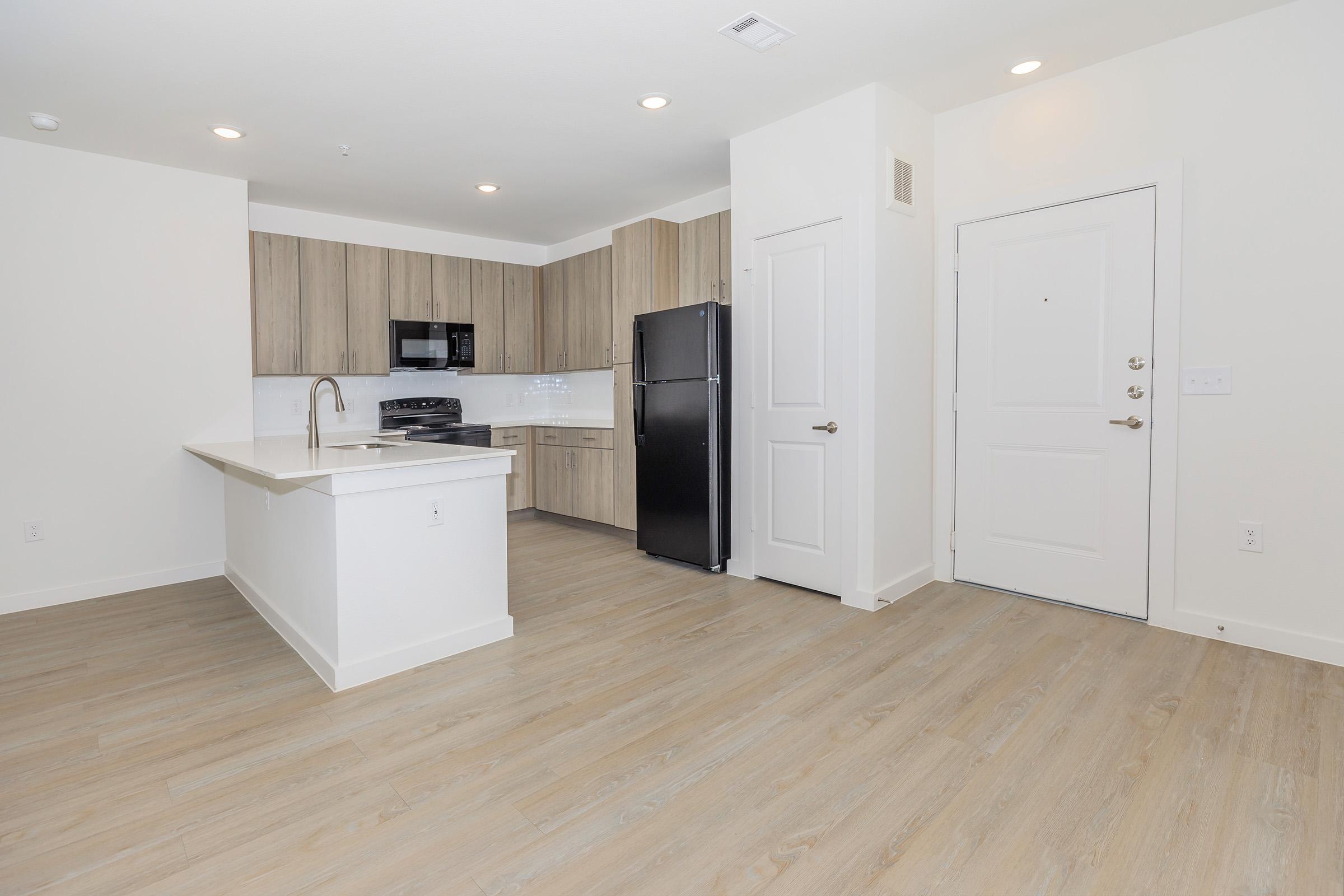 Modern kitchen and living area featuring light wood flooring, granite countertops, and stainless steel appliances. The kitchen is equipped with a black refrigerator, oven, and microwave. A front door is visible, leading to a well-lit and spacious environment.