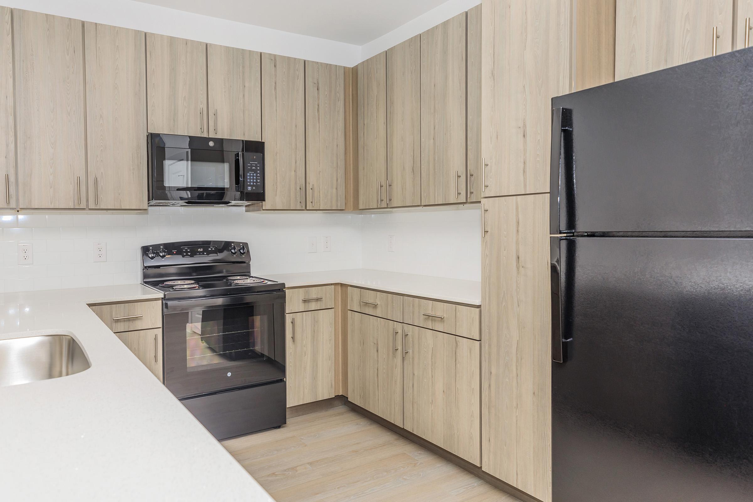 Modern kitchen featuring light wood cabinets, stainless steel appliances, including a microwave and oven, and a black refrigerator. The countertop is white, and the flooring is light-colored wood. The space appears clean and well-organized, ideal for cooking and dining.