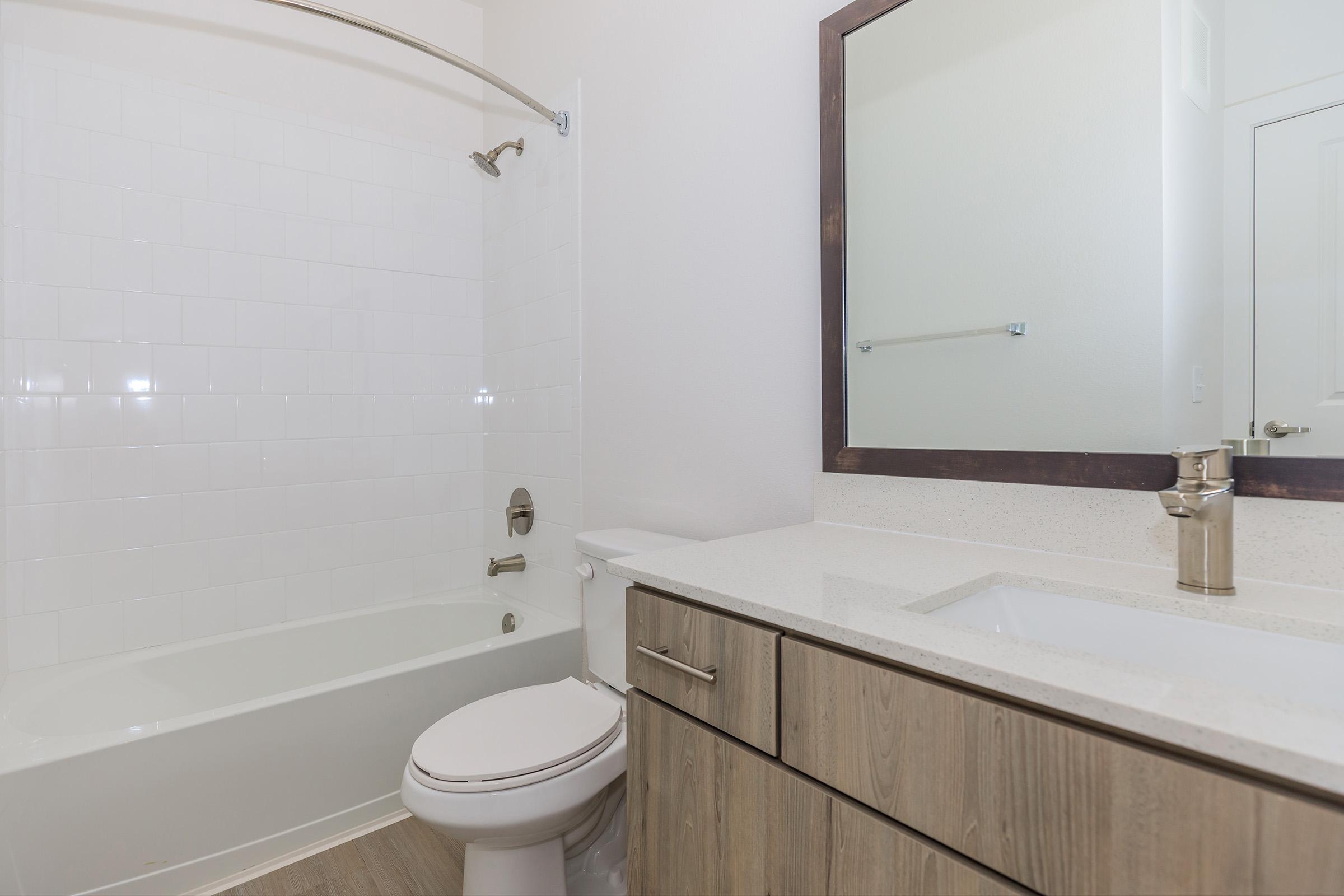A modern bathroom featuring a shower-tub combination with white tiles, a light wood vanity with a sink, a large mirror, and a sleek faucet. The floor is a light wood pattern, creating a clean and contemporary look.