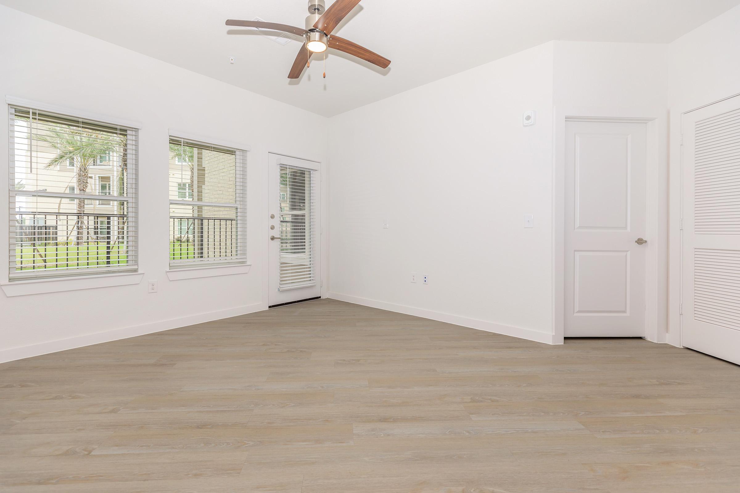 Bright and airy interior of a room featuring light wooden flooring, large windows with blinds, and a ceiling fan. There's a sliding glass door leading to an outdoor view, and a closed door to the right. The walls are painted white, creating a clean and modern atmosphere.