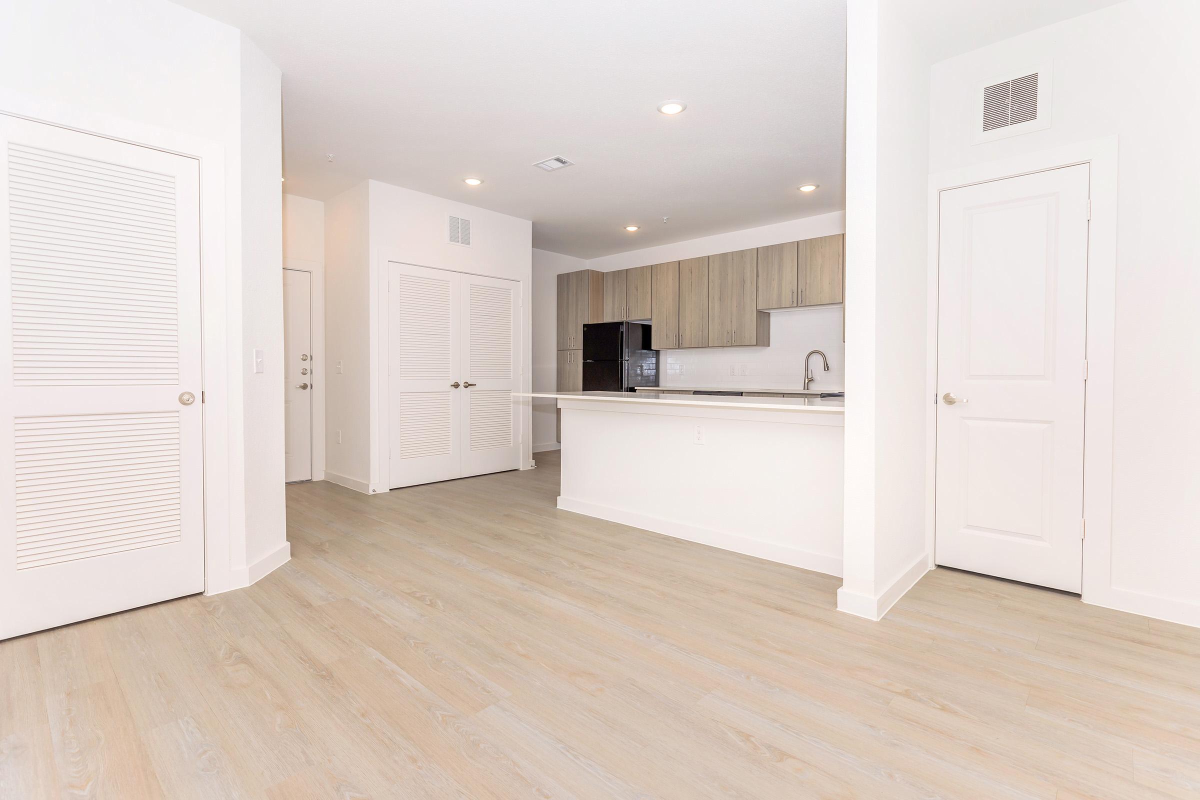 Bright and modern kitchen and living space featuring light-colored wood flooring, white walls, and contemporary cabinetry. The open layout includes a kitchen with a black refrigerator and sleek counter space, with two sets of white doors leading to adjacent rooms. Ample natural light illuminates the area.