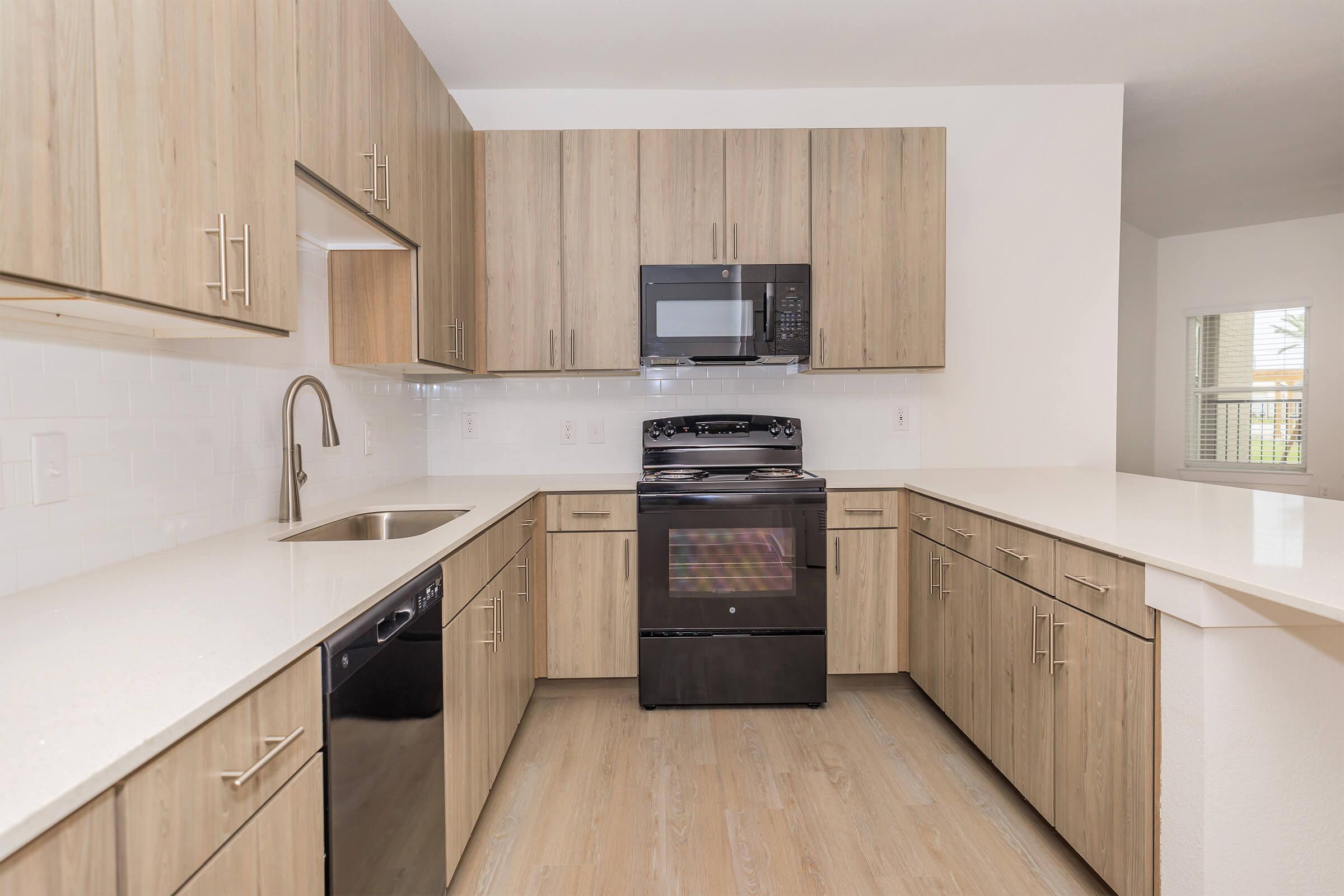 Modern kitchen with light wood cabinetry, stainless steel appliances including a microwave and stove, a black dishwasher, and a spacious countertop. The design features a clean, minimalist style with ample storage and natural light coming through a nearby window.