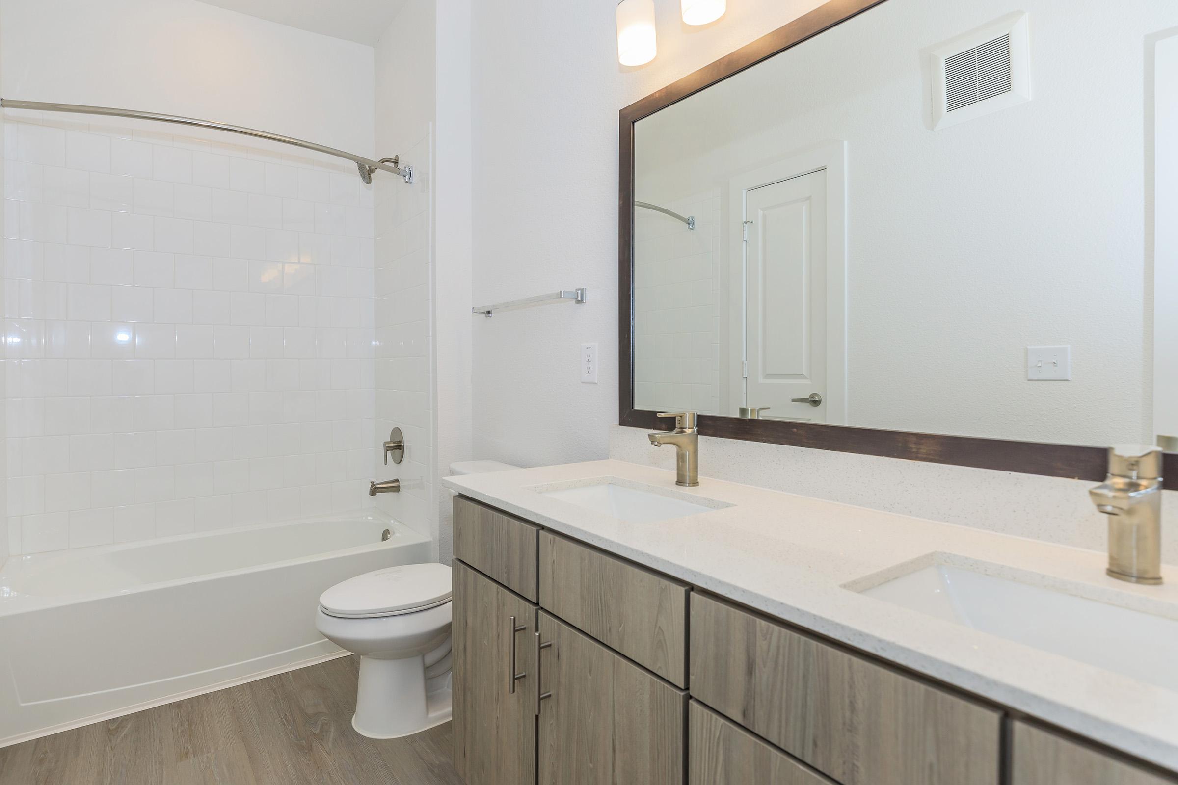 Modern bathroom featuring a bathtub with a white tiled wall, a double vanity with two sinks, light wood cabinetry, and stylish gold faucets. There is a large mirror above the sinks and soft lighting fixtures on the wall, offering a clean and contemporary aesthetic.