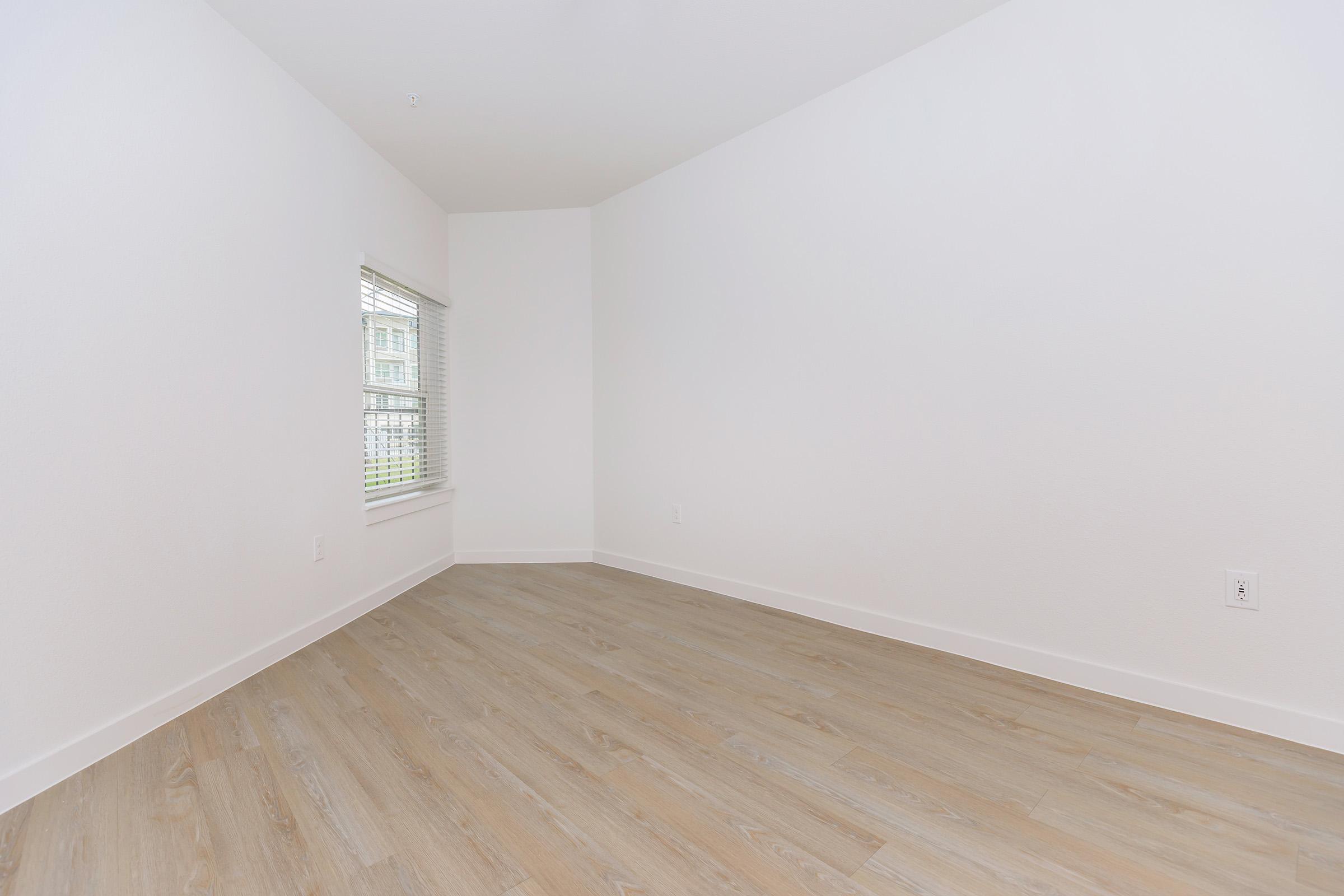 Empty room with light-colored walls, a large window covered by blinds, and light wooden flooring. The space is bright and minimalistic, with no furniture or decorations visible.