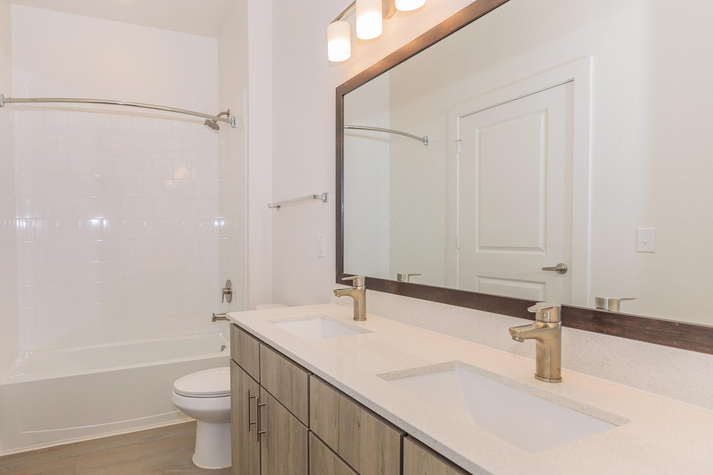 A modern bathroom with a white tiled bathtub and shower, a long vanity with double sinks, stylish gold faucets, and a large rectangular mirror. The walls are painted in a light color, complemented by natural wood cabinetry. Bright lighting fixtures are mounted above the mirror.