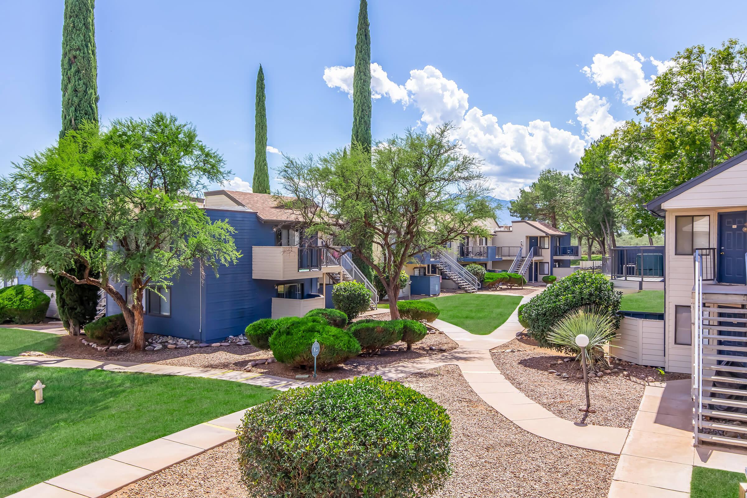 A well-maintained residential complex featuring multiple buildings with blue exteriors, surrounded by neatly trimmed lawns, landscaped shrubs, and tall trees. The scene includes pathways connecting the buildings under a bright blue sky with fluffy clouds.