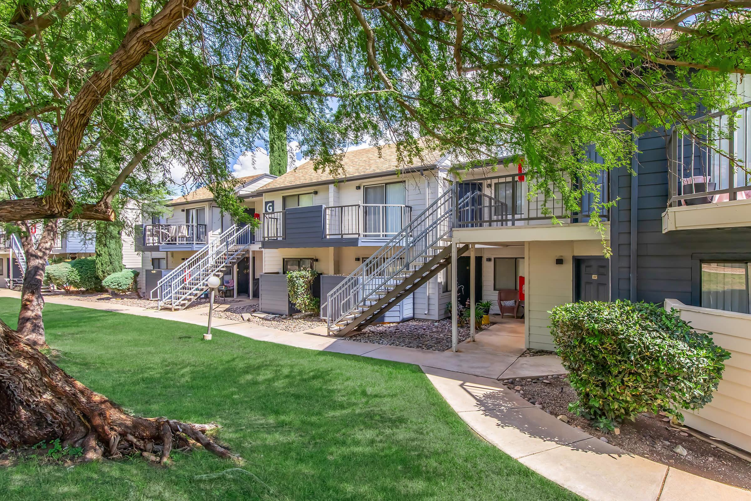 A landscaped courtyard featuring several apartment buildings with stairs leading to individual units. Lush green grass and a large tree provide shade, while walkways connect the apartments. The buildings have balconies and varied exterior colors, showcasing a pleasant residential environment.