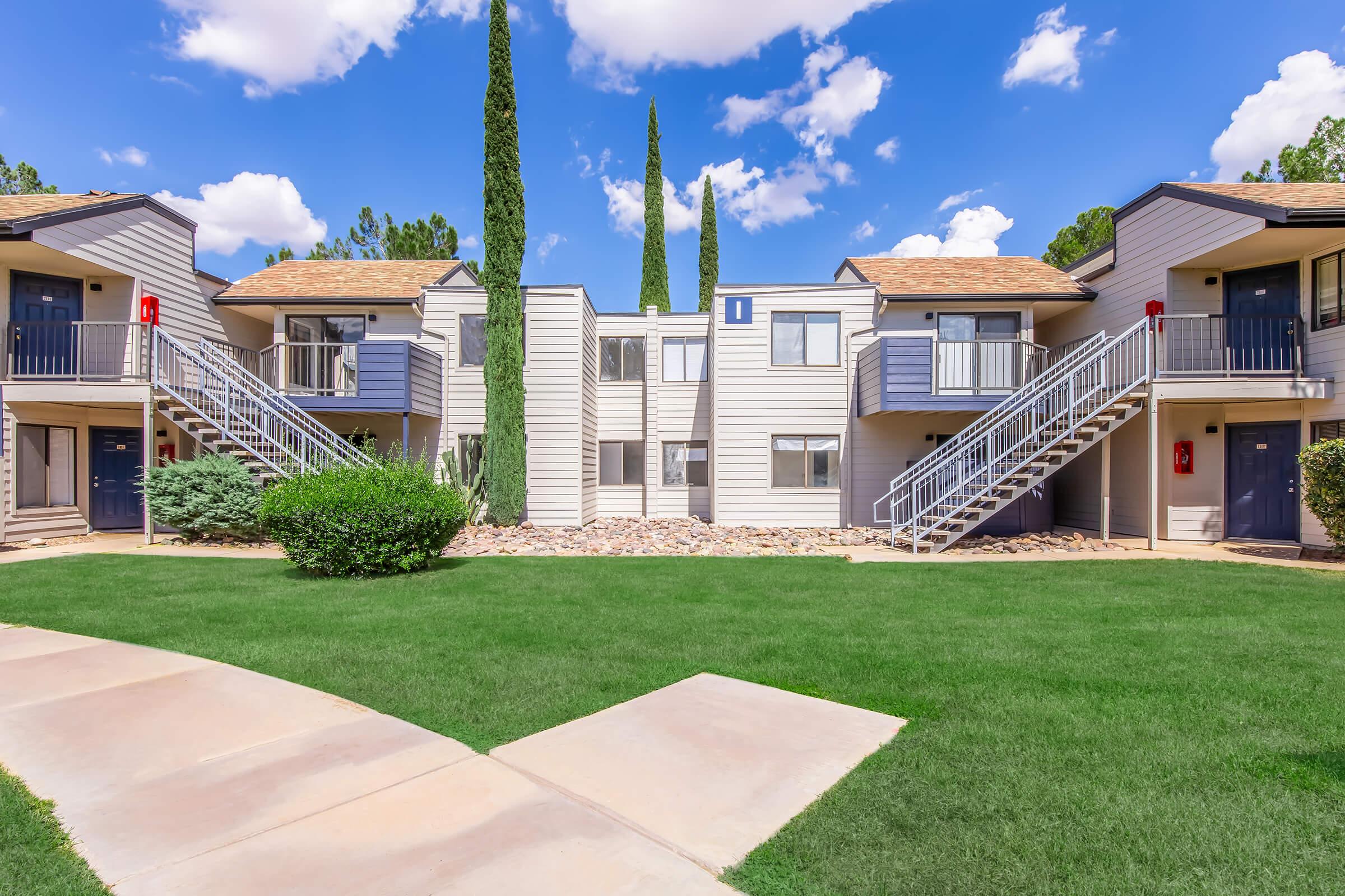 A view of a residential complex featuring two-story buildings arranged around a central green lawn. The buildings have balconies and staircases, with tall cypress trees in the background and blue sky with fluffy white clouds overhead. Pathways lead through the grassy area.