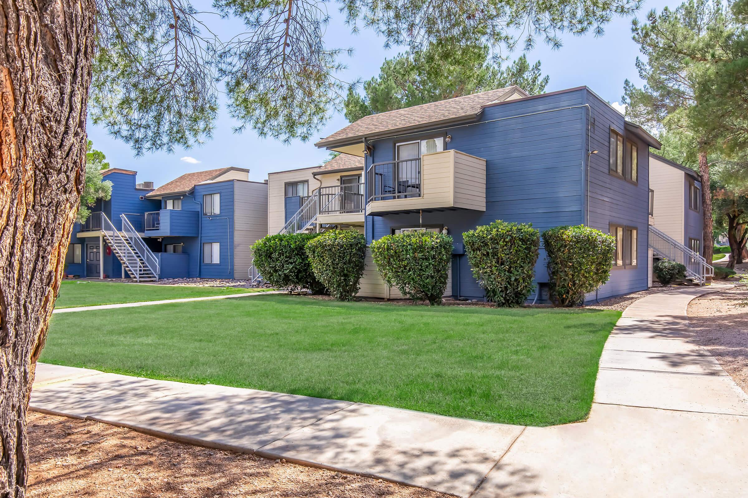 Two-level apartment buildings with blue exteriors and balconies, surrounded by well-manicured green lawns and neatly trimmed hedges. A clear pathway runs alongside the buildings, and trees provide shade in the background under a sunny sky.